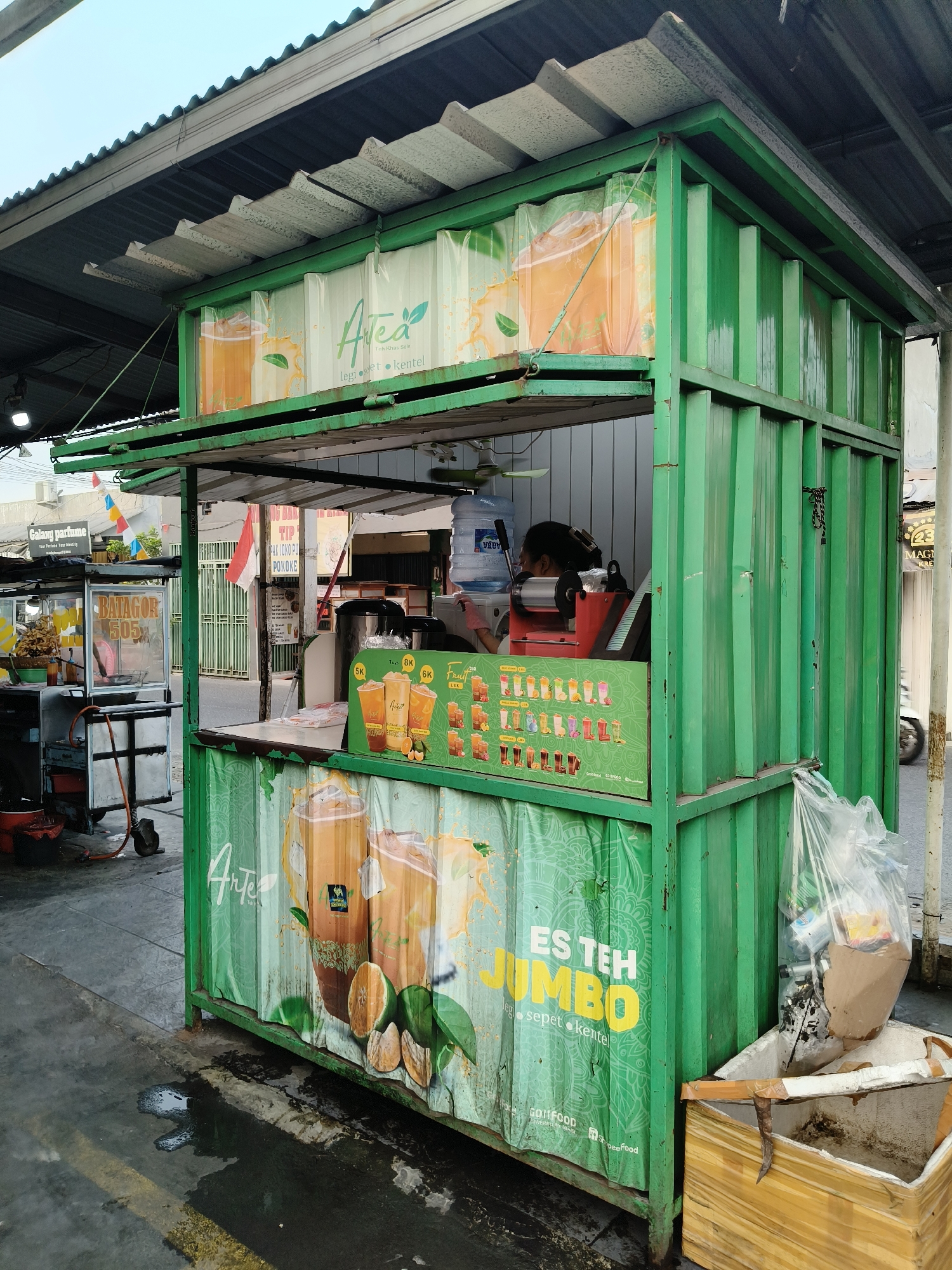 This image depicts an outdoor street food stall, specifically a "JUMBO ES TEH" (Jumbo Iced Tea) stand, likely located in South Tangerang, Indonesia, given the context of local street food culture. The stall itself is constructed from a green shipping container, painted a vibrant green. It features a prominent banner with images of iced tea drinks and the text "JUMBO ES TEH". A smaller banner displays various drink options with prices in Indonesian Rupiah.
Inside the stall, a woman with dark hair is visible behind the counter, attending to operations. Equipment such as a water dispenser and a sealing machine for drinks are present. To the left of the main stall, another food cart is visible, possibly selling fried snacks, indicated by a sign reading "BATAGOR 505".
The ground in front of the stall is wet and appears to be paved, with some yellow lines painted on it, suggesting a street or walkway. A large, clear plastic bag filled with trash is seen to the right of the stall, next to a cardboard and wooden receptacle for waste. In the background, other street vendors and buildings are visible, with a motorcycle passing by. The overall scene captures a typical roadside food vending environment in an urban Indonesian setting.