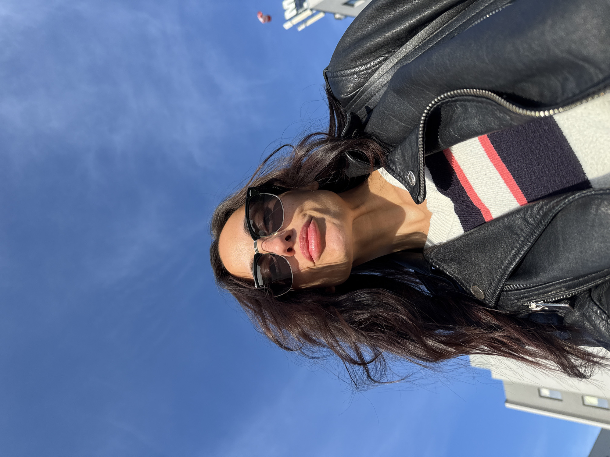 A selfie-style, upward-angled shot features a woman wearing sunglasses and a black leather jacket. A white sweater with a striped collar is visible beneath the jacket. The background is a clear blue sky with faint cloud streaks. In the upper portion of the image, the top of a building is visible. It is not possible to ascertain the location in Pleven, Bulgaria.