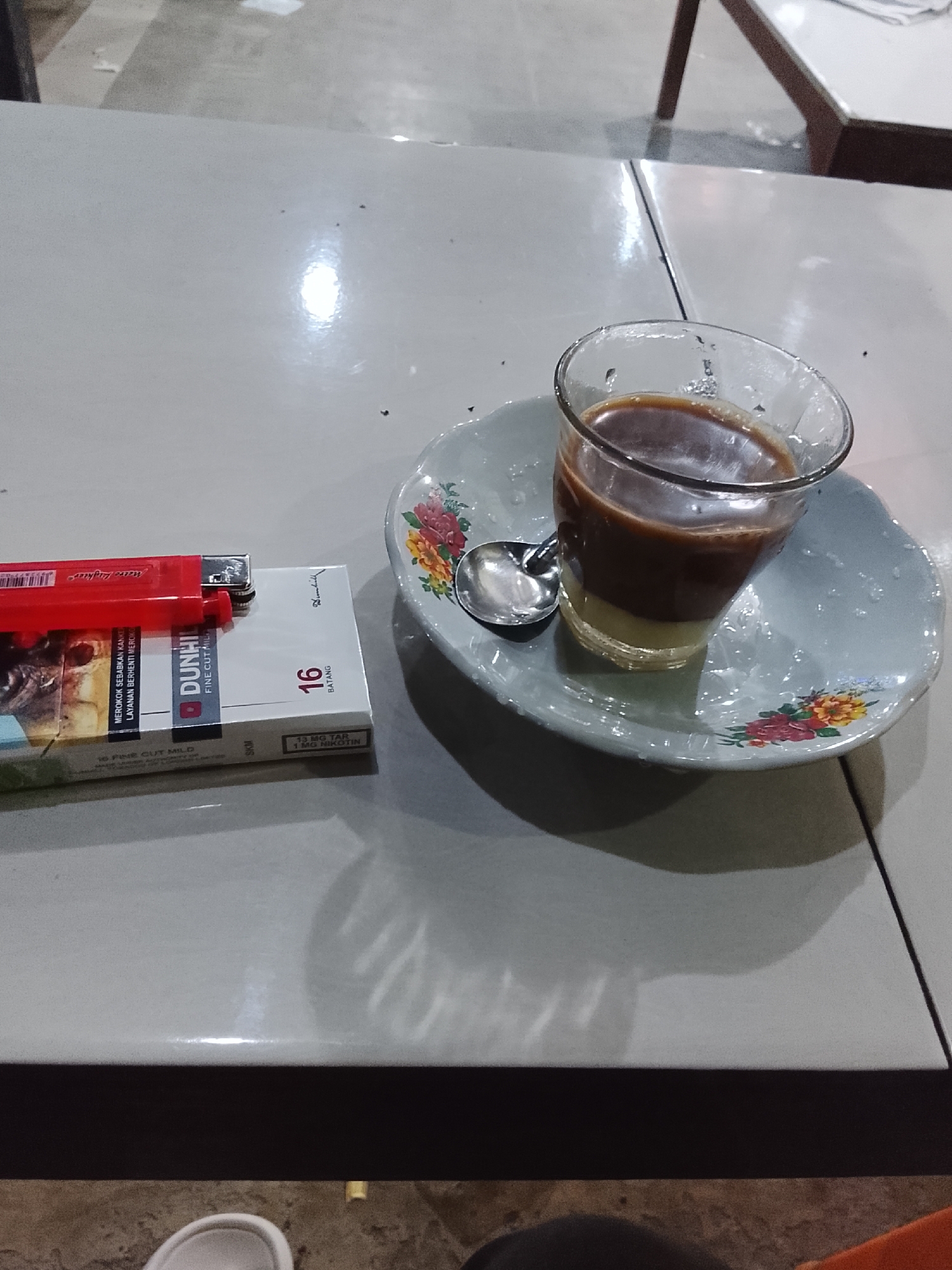 A red lighter rests on a pack of Dunhill Fine Cut Mild cigarettes. To the right, a glass of coffee with condensed milk sits on a floral saucer next to a spoon. The scene is set on a white, possibly plastic, table. The background is blurred, showing hints of a seating area with a dark wooden table. The overall environment suggests a casual outdoor or indoor setting, possibly a cafe or roadside stall, within Dayahlhoksukon Barat, Indonesia.