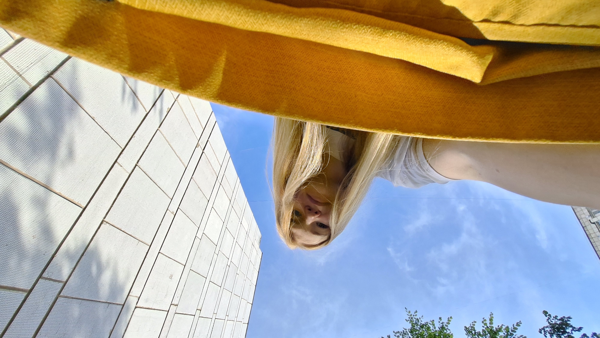 A low-angle selfie shows a young woman with long blonde hair looking down towards the camera, her face framed by the bright yellow fabric of what appears to be a blanket or garment in the foreground. She is wearing a white t-shirt. Behind her, a clear blue sky with wispy clouds is visible, along with the upper branches of a leafy tree on the left. To the right, a modern building with a facade of light-colored, rectangular tiles rises sharply against the sky. The overall impression is of an outdoor, urban setting. This scene could be in the city of Chaikovsky, Russia, given the architectural style of the building and the commonality of such urban landscapes in Russia.