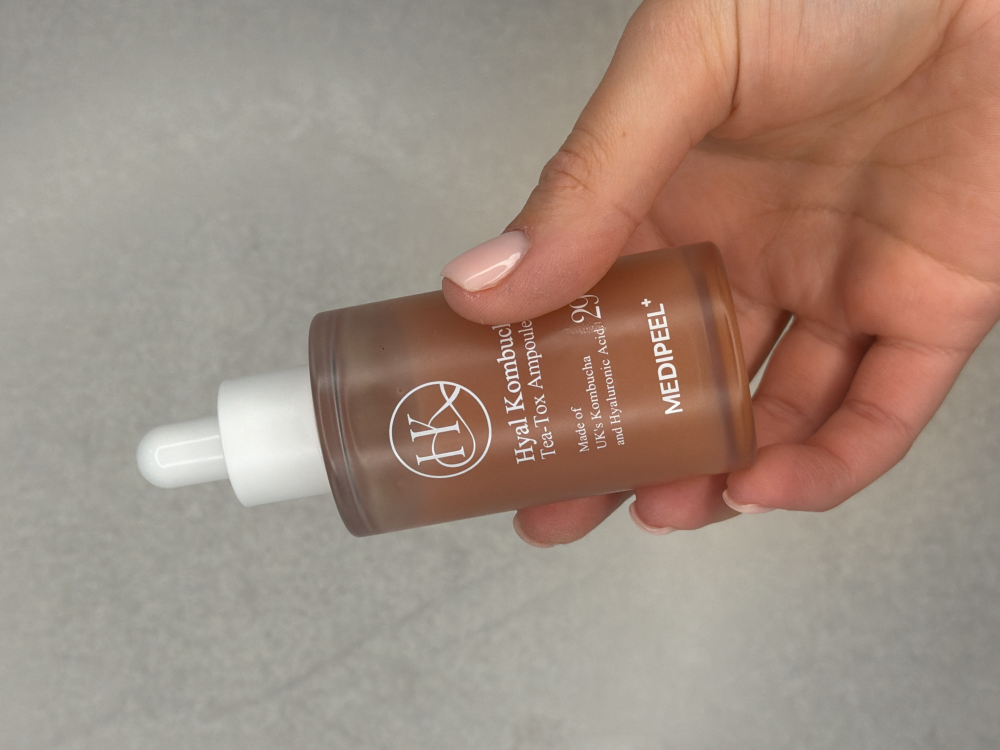 An eye-level, close-up shot features a person's hand holding a cosmetic ampoule bottle against a neutral, light grey textured background. The hand, with manicured nails painted in a subtle nude-pink color, holds the amber-tinted glass bottle horizontally. The bottle is equipped with a white dropper top.
The visible text on the bottle identifies the brand as "MEDIPEEL+" and the product as "Hyal Kombuc Tea-Tox Ampoule." It also specifies that the product is "Made of UK's Kombucha and Hyaluronic Acid." A numerical value "29" is present, along with a circular logo featuring the letter "(K)". The image appears to be a product shot, brightly and evenly lit. The background is simple, ensuring focus remains on the product and the hand holding it. The setting appears to be indoors, possibly in Sofiivska Borshchahivka, Ukraine, as suggested by the external context, but no specific location cues are visible within the image itself.