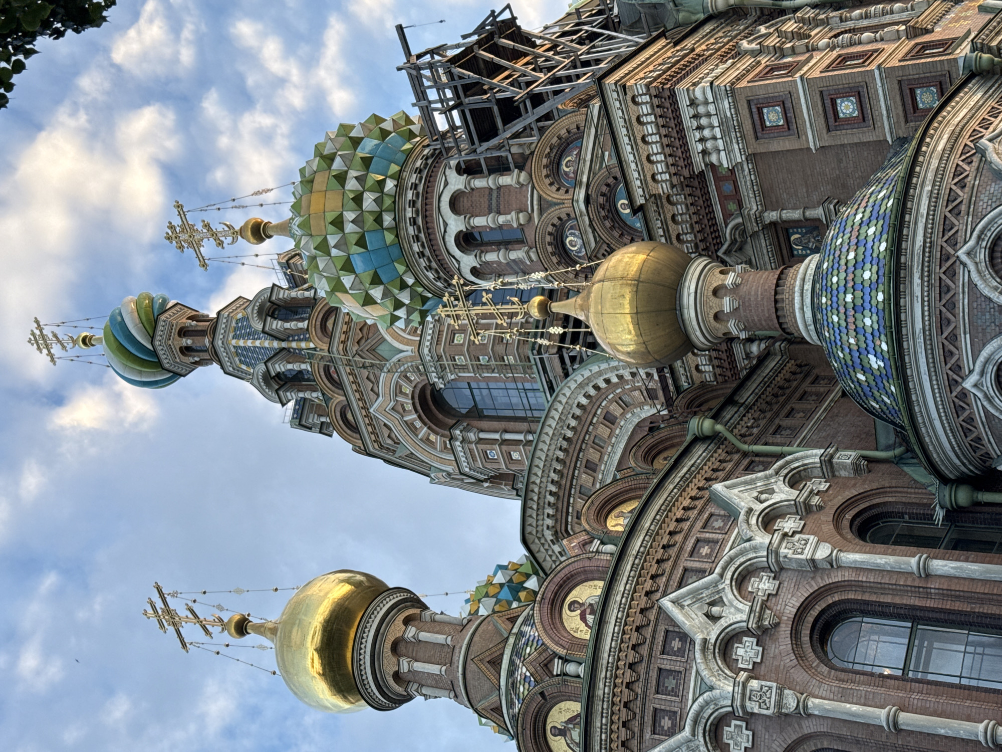 The image captures a detailed, low-angle view of the Church of the Savior on Spilled Blood in Saint Petersburg, Russia. The iconic onion domes, one prominent and golden, and another smaller, mosaic-tiled in shades of blue, green, and white, are visible. The church's ornate facade, constructed from red brick with intricate white detailing and mosaic inlays depicting religious figures, is a central focus. Several crosses, characteristic of Orthodox churches, adorn the domes. The sky above is partly cloudy with a light blue hue. Scaffolding is visible on the right side of the church, suggesting ongoing restoration or maintenance work. The lighting indicates it is daytime, possibly late afternoon, with sunlight casting soft shadows. No people are present in the frame.