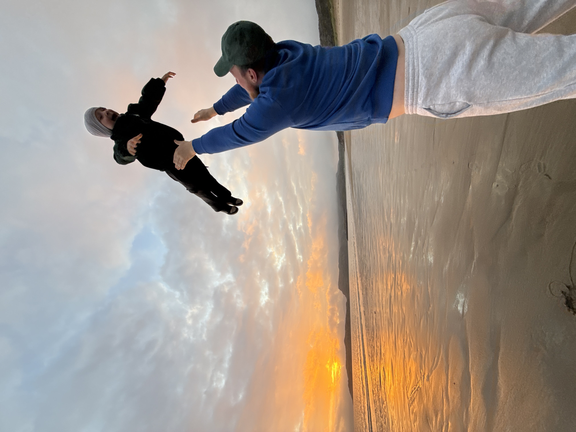 A man in a blue hoodie and grey sweatpants throws a child in a black jacket and grey beanie high into the air on a beach at sunset. The sky is filled with clouds that are illuminated by the orange glow of the setting sun. The ocean reflects the colors of the sky, and the wet sand on the beach also reflects the light. In the distance, there are hills and mountains. The scene is located on a beach in Teriberka, Russia.