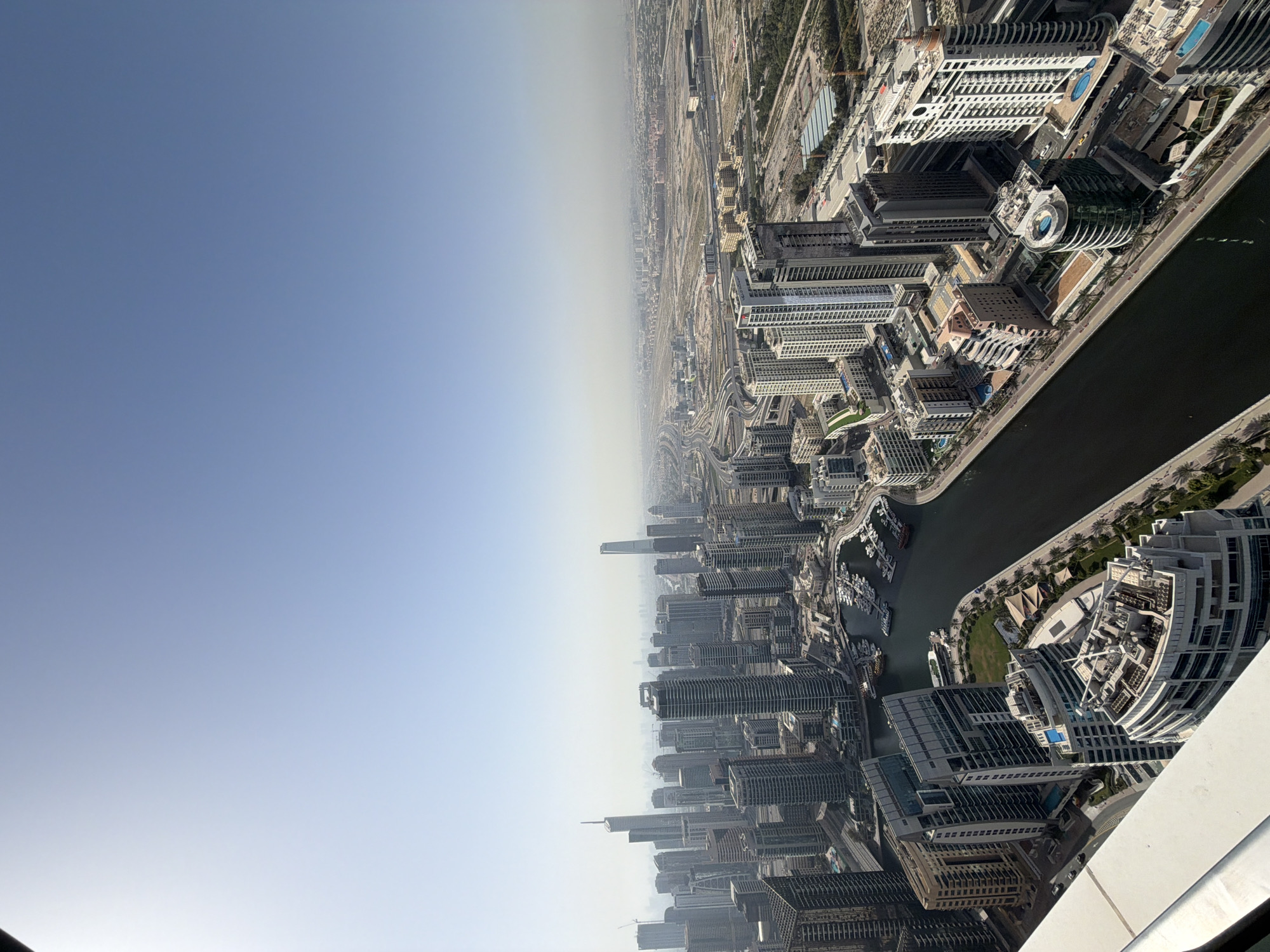 The image presents an aerial view of Dubai Marina in the United Arab Emirates. A waterway bisects the urban landscape, flanked by high-rise buildings and recreational areas. The sky above transitions from a deep blue to a hazy horizon.