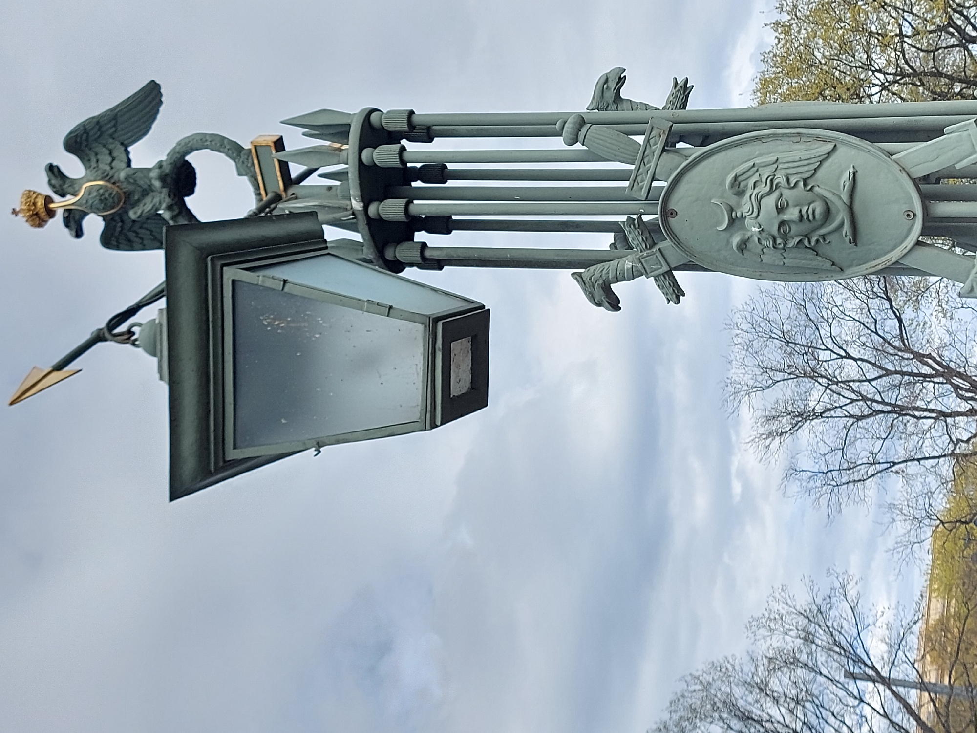 The image shows a street lamp in Saint Petersburg, Russia. The lamp has a grey-green ornate design, including a double-headed eagle with a gold crown, a lantern, and a relief of a face with wings. The lamp is set against a cloudy sky and bare tree branches.