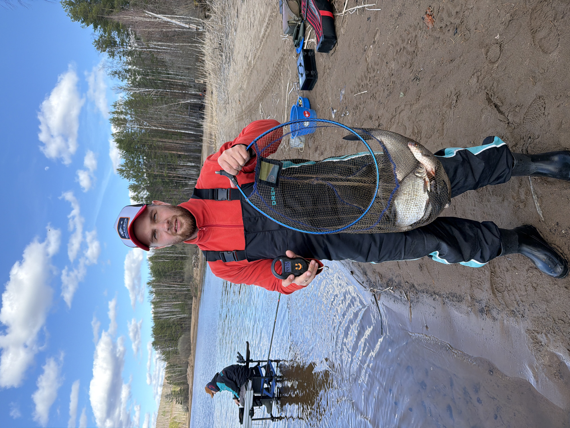 A man stands on a sandy shore, holding a blue fishing net filled with fish. He is wearing a red jacket and black trousers with a light blue stripe, as well as black rubber boots and a baseball cap. The man is also holding a digital scale in his hand. Behind him is a lake and a forest. Another person is fishing in the lake using a fishing platform. The sky is blue with scattered clouds. The location appears to be Chaykovsky, Russia.