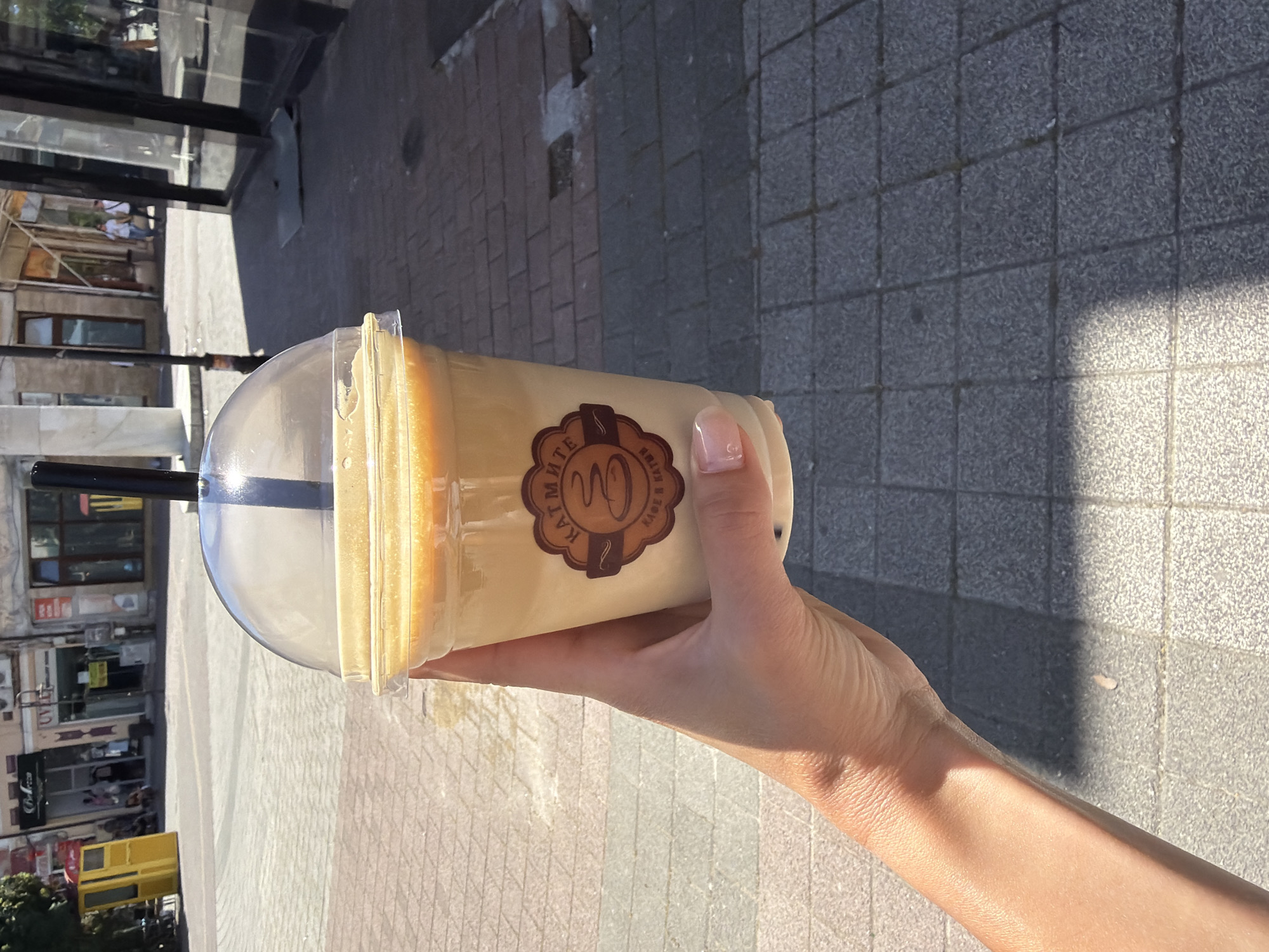 A hand holds a clear plastic cup filled with a light brown beverage, likely coffee or tea, topped with a domed lid and a black straw. The cup has a circular brown logo with text that appears to read "КАТМИ" and "КАФЕ & NATUR". The background shows a sunlit paved sidewalk and a street in the city of Pazardzhik, Bulgaria. Buildings with storefronts are visible across the street, some featuring yellow and brown signage. A yellow kiosk-like structure is also present. The paving stones are laid in a grid pattern, with some areas casting shadows. The overall scene suggests a casual outdoor setting during the daytime.