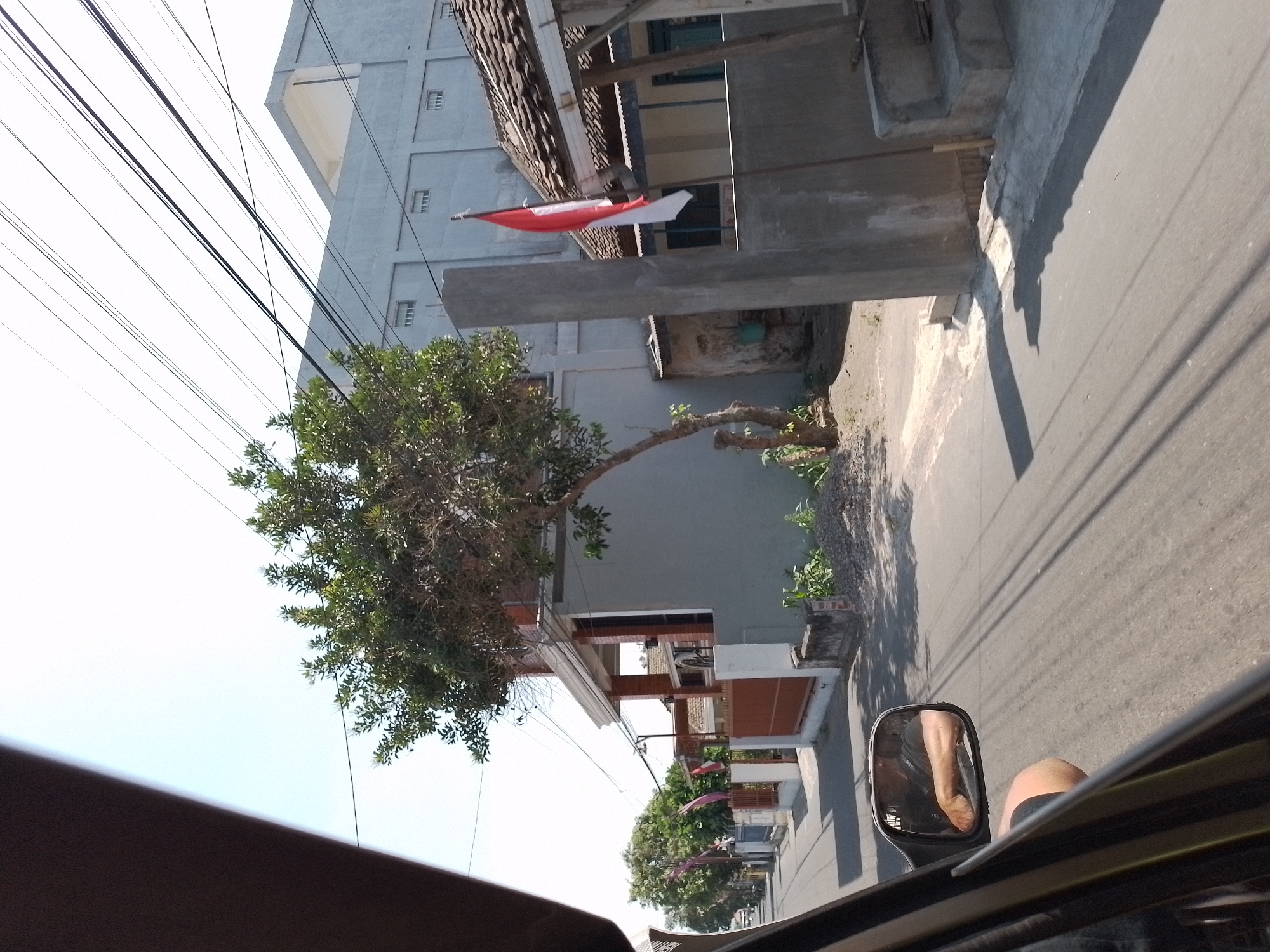 The image captures a street scene in Wates, Indonesia, on a bright, sunny day. The perspective is from inside a vehicle, looking out the side window and at the side mirror. A person's arm, wearing a dark t-shirt and shorts, is visible in the side mirror, resting on the car door. The street is paved and runs alongside houses and buildings. On the right side of the street, a prominent Indonesian flag, red and white, is mounted on a pole near a building with a tiled roof. To the left of the flag, a tall, gray concrete pillar stands. Behind the flag and pillar, a large, two-story building with a gray facade and horizontal windows is visible. Further down the street on the left, a smaller, two-story house with a gray exterior and red-brown gates is present, surrounded by trees and greenery. Another building, also with a gray facade, is visible in the distance, along with more trees and what appear to be smaller houses or structures. Utility wires are strung across the sky. The overall impression is a typical residential street in a town, likely during a national celebration given the prominent display of the Indonesian flag.