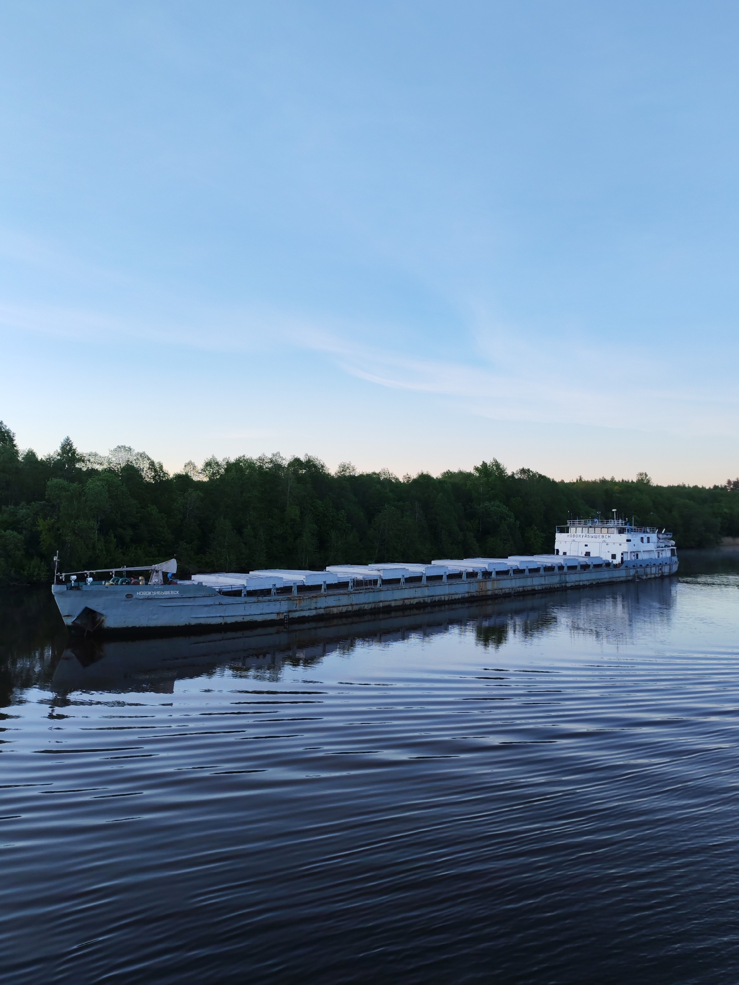 This media file captures a long, grey cargo barge moving or moored on a calm river. The barge has numerous large, light-colored covers over its cargo holds and a multi-deck white cabin structure at its stern. Dense green forest lines the distant riverbank, creating a natural backdrop.
The sky is a clear, light blue with subtle wisps of clouds, suggesting either early morning or late evening due to the soft, even light. The river's surface is smooth with gentle ripples, reflecting the sky and the trees. The overall atmosphere is peaceful and serene.
Visible white text on the bow of the barge, as well as on the side of the stern cabin, reads "НОВОКУЙБЫШЕВСК" (Novokuybyshevsk), likely indicating its name or homeport. No people are visible in the image. The setting appears to be a natural waterway, consistent with the location of Volokov Most, Russia.