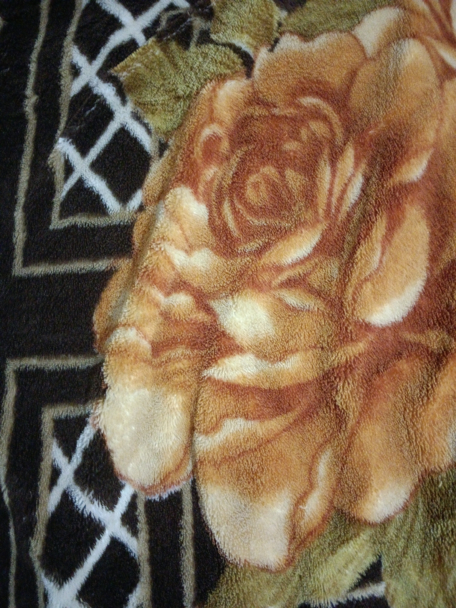 The image shows a close-up view of a fuzzy blanket with a floral pattern. A large, textured rose in shades of orange, brown, and cream dominates the center of the frame. The rose appears to be rendered in a plush, velvety material, with distinct petal formations. To the left of the rose, a darker background features a geometric pattern of interlocking white lines that form a diamond or lattice design against a black base. The overall impression is one of tactile softness and a rich, somewhat abstract floral motif. There is no discernible location in the city of Karapalli, India, or any people, subjects, actions, or interactions depicted.
