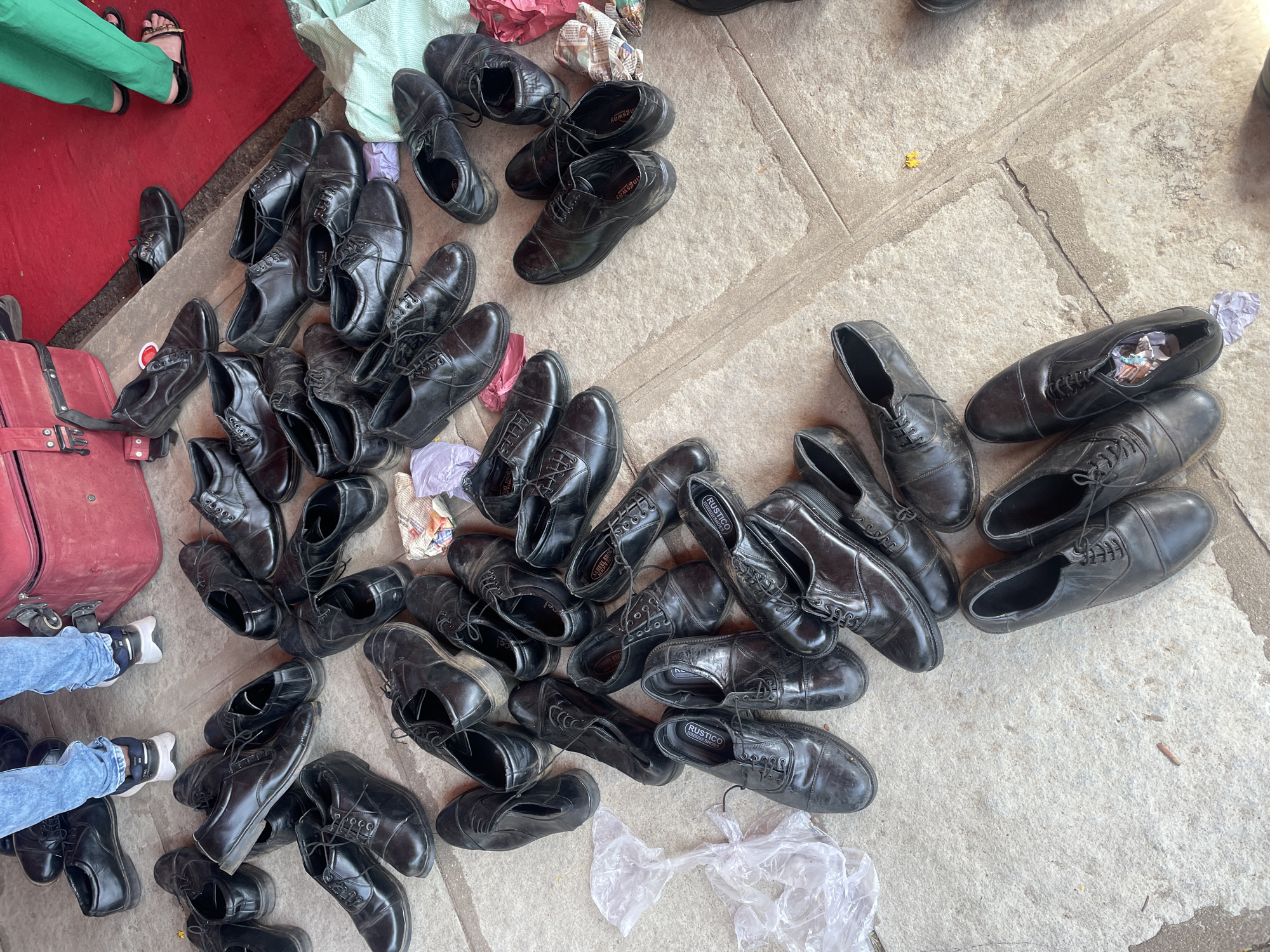 A large pile of black leather dress shoes is scattered on a concrete surface. Some shoes contain crumpled paper or tissue inside. A red suitcase is visible to the left, partially covered with shoes. Two people's legs are seen; one wearing green pants and sandals, the other wearing jeans and sneakers. The shoes appear worn, suggesting they are used or for sale. The scene, likely a marketplace or street vending area, possibly takes place in Anajpur, India.