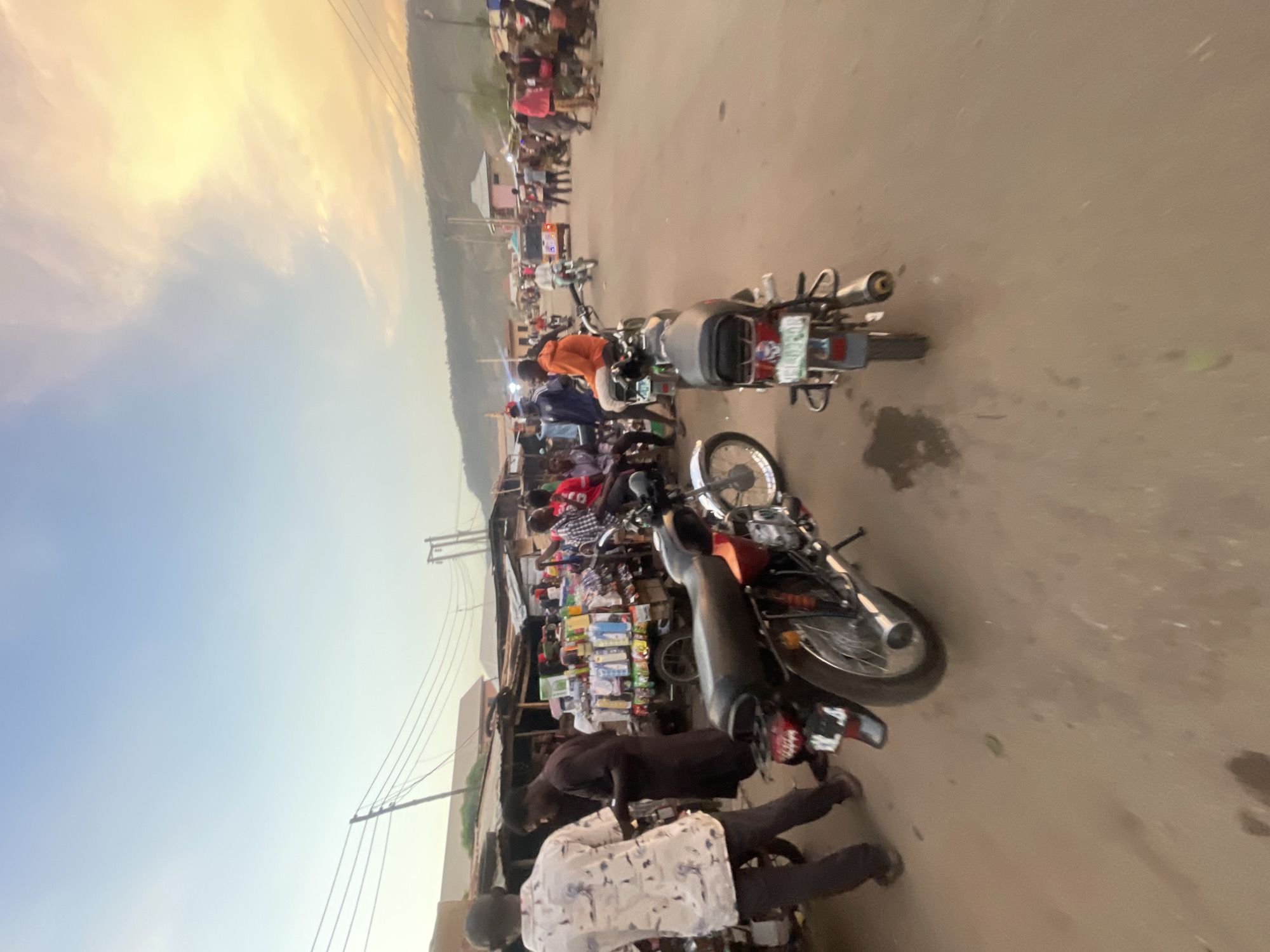 The image shows a street scene in Lokoja, Nigeria, likely near a market area. The shot is taken at ground level, angled to the side. The sky is visible in the background, showing either sunrise or sunset. Several motorcycles are parked on the right side of the road, partially blocking the street. A row of small shops or stalls line the left side of the street. People are milling about, interacting with the vendors or simply passing through. The atmosphere is busy.