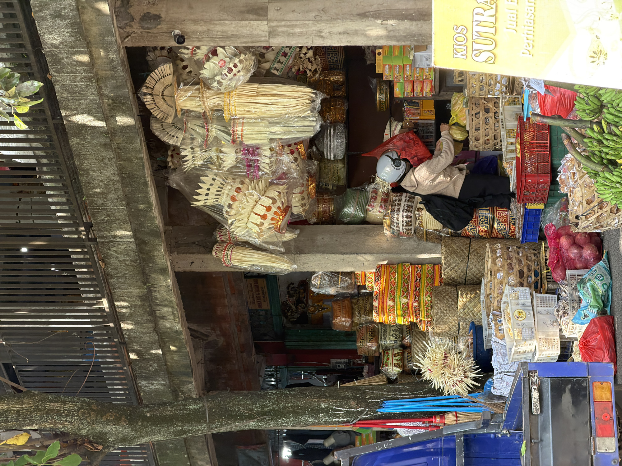 The image shows a street scene in the Ubud District, Indonesia, specifically in front of a shop selling traditional crafts and produce. A woman wearing a light-colored jacket, dark pants, and a white helmet is visible in the center of the frame. She is standing in front of a stall, seemingly interacting with the goods, with her back partially to the camera.
The shop is filled with various items. Hanging from above are numerous woven decorative items, many wrapped in plastic. Below these, the shelves and surfaces are stacked with intricately woven baskets and containers of different sizes and patterns, featuring geometric designs in yellow, red, green, and brown. There are also piles of stacked boxes and some produce, including red onions in a red mesh bag and bunches of green bananas in the foreground.
To the left of the stall, a blue vehicle with a black cargo bed is parked. A broom with red and blue handles is leaning against it. A large tree with textured bark stands to the far left of the image, partially obscuring the background. The overall impression is a vibrant marketplace scene with a focus on artisanal goods. A yellow sign with the text "KIOS SUTRA" and other Indonesian writing is prominently displayed on the right side of the image. The lighting suggests it is daytime, with sunlight casting shadows.