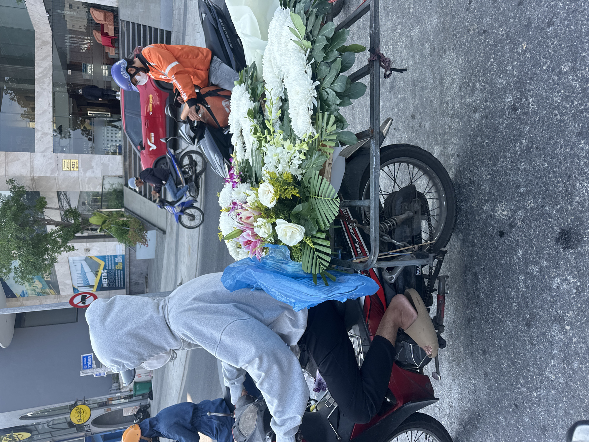 The image shows a motorcycle heavily loaded with a large bouquet of white and colorful flowers secured to the back carrier. The driver, wearing a gray hooded sweatshirt, black pants, and beige sandals, sits on the red motorcycle.
In the background, a man in an orange jacket and helmet sits on another motorcycle. Other motorcycles are parked nearby. Buildings are visible, including one with a sign that reads "MEETING ROOM" and "532." Street signs indicate the location is in Đà Nẵng, Vietnam: "BƯỜNG 2 THÁNG 9". The presence of "ShopeeFood" on the orange jacket suggests delivery service. The overall scene depicts a flower delivery or transport in an urban setting in Đà Nẵng.