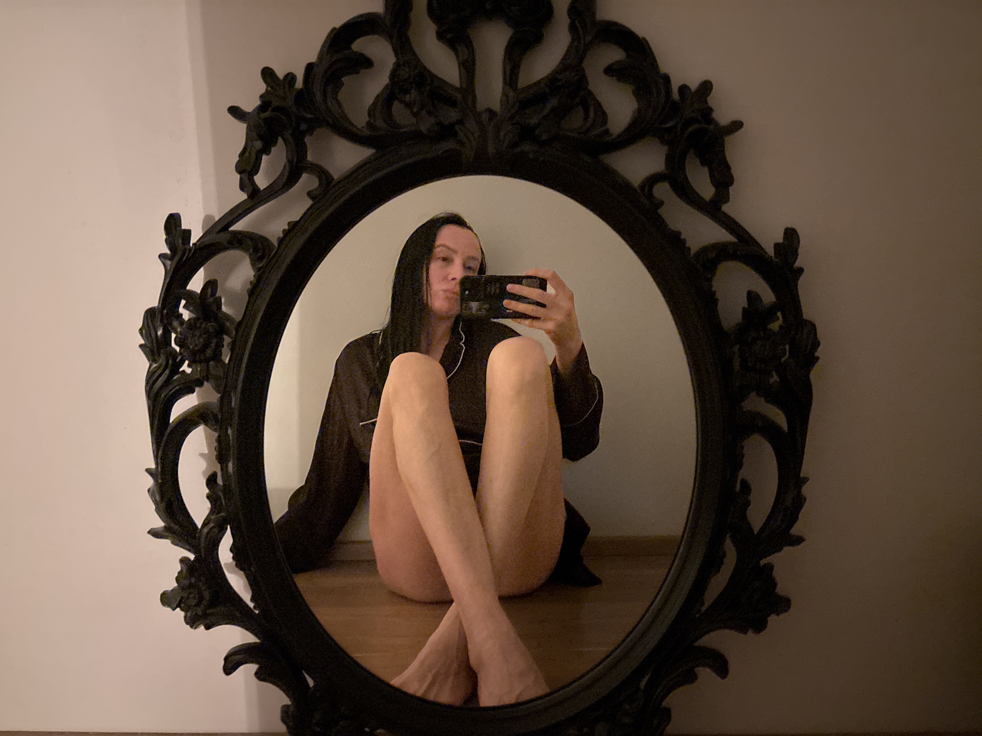 A photograph shows a person reflected in a large, ornate, oval-shaped black mirror. The person is seated on the floor, with their legs crossed. They are wearing a long-sleeved, dark-colored pajama top with white trim and appear to be taking a selfie with a smartphone. The mirror's frame is highly decorative, featuring floral and foliate motifs. The wall and floor behind the mirror appear to be plain and light in color. The lighting suggests an indoor setting, likely a home. The location is Chaikovsky, Russia.
