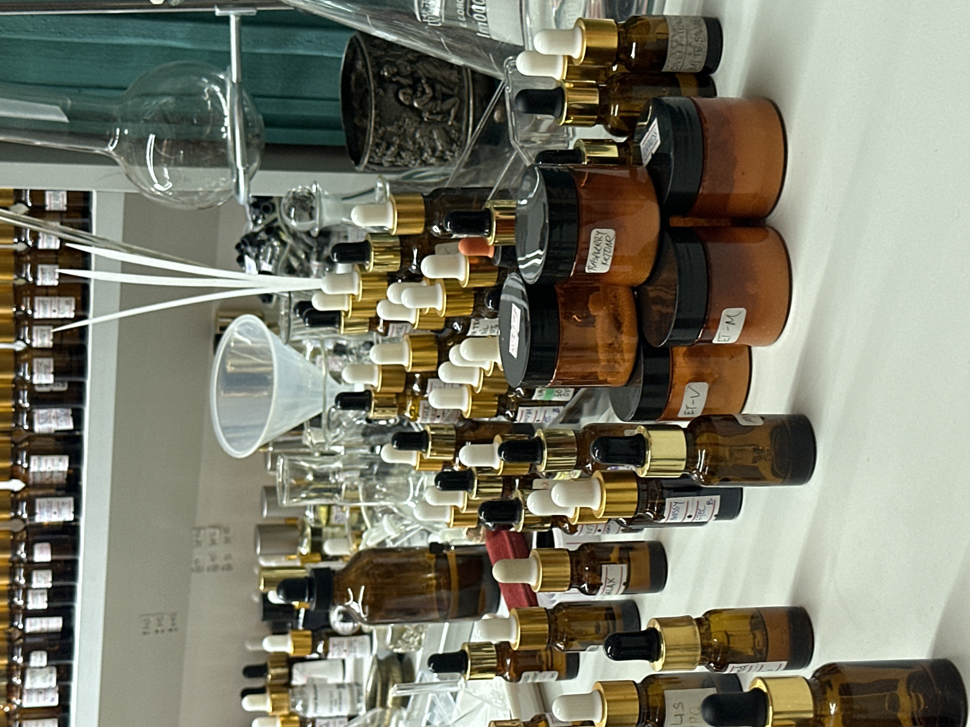 An indoor, eye-level, close-up shot shows a collection of small amber glass bottles with dropper lids and several small amber jars filled with an orange-colored substance. The bottles and jars are arranged on a white surface, with more bottles visible in the background on shelves. Some bottles have labels with handwritten text, such as "LAX," "ET-V," and "RASPBERRY KETONE." In the background, there are laboratory glassware, including a funnel and flasks. The scene appears to be a workshop or laboratory.