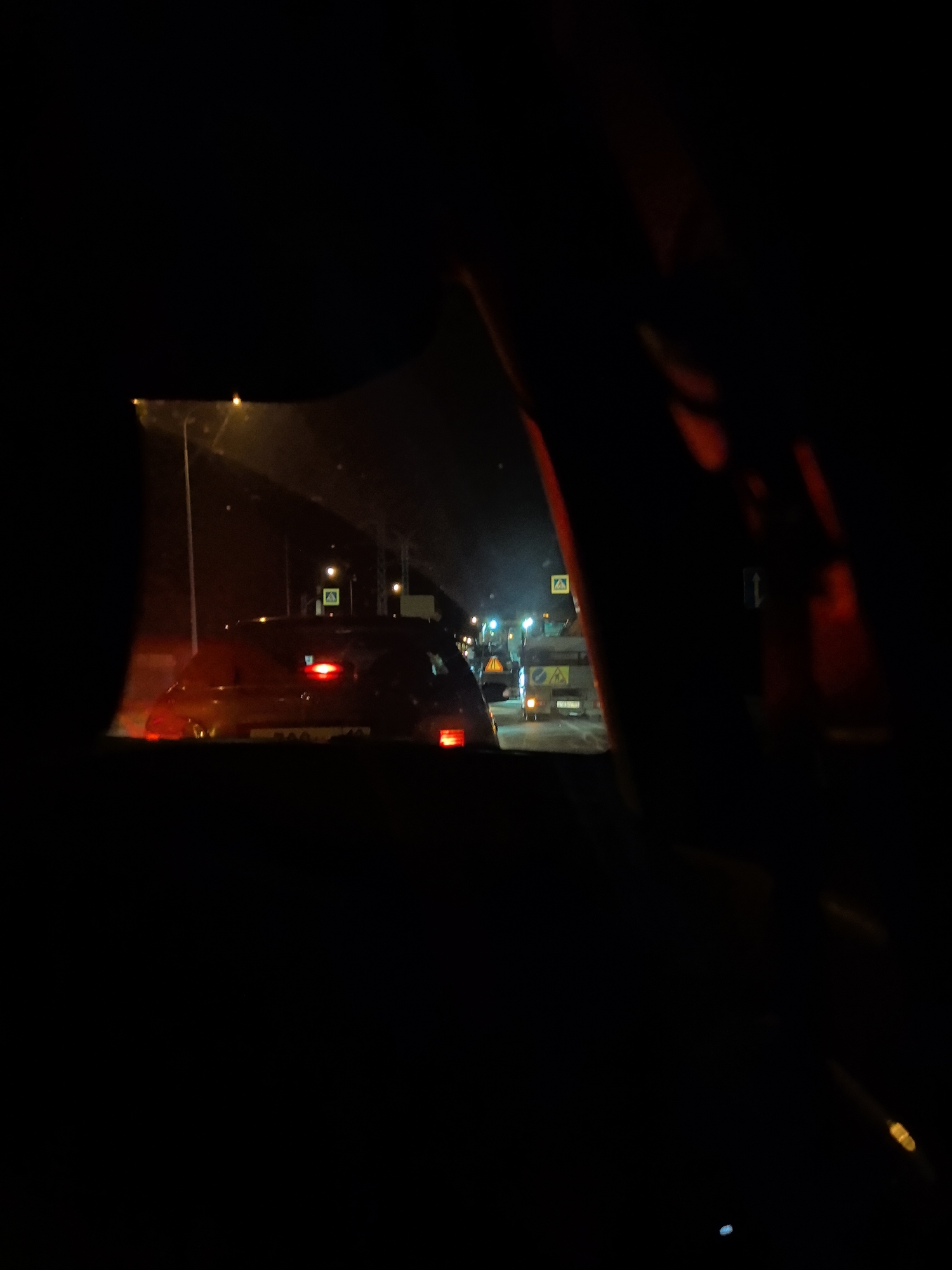 The image shows a night scene from inside a vehicle. Several cars are visible ahead, with illuminated taillights. A street light is visible on the left and a utility truck with warning signs is further ahead in the traffic. Yellow road work signs can be seen in the distance. The location is in Chaikovsky, Russia.