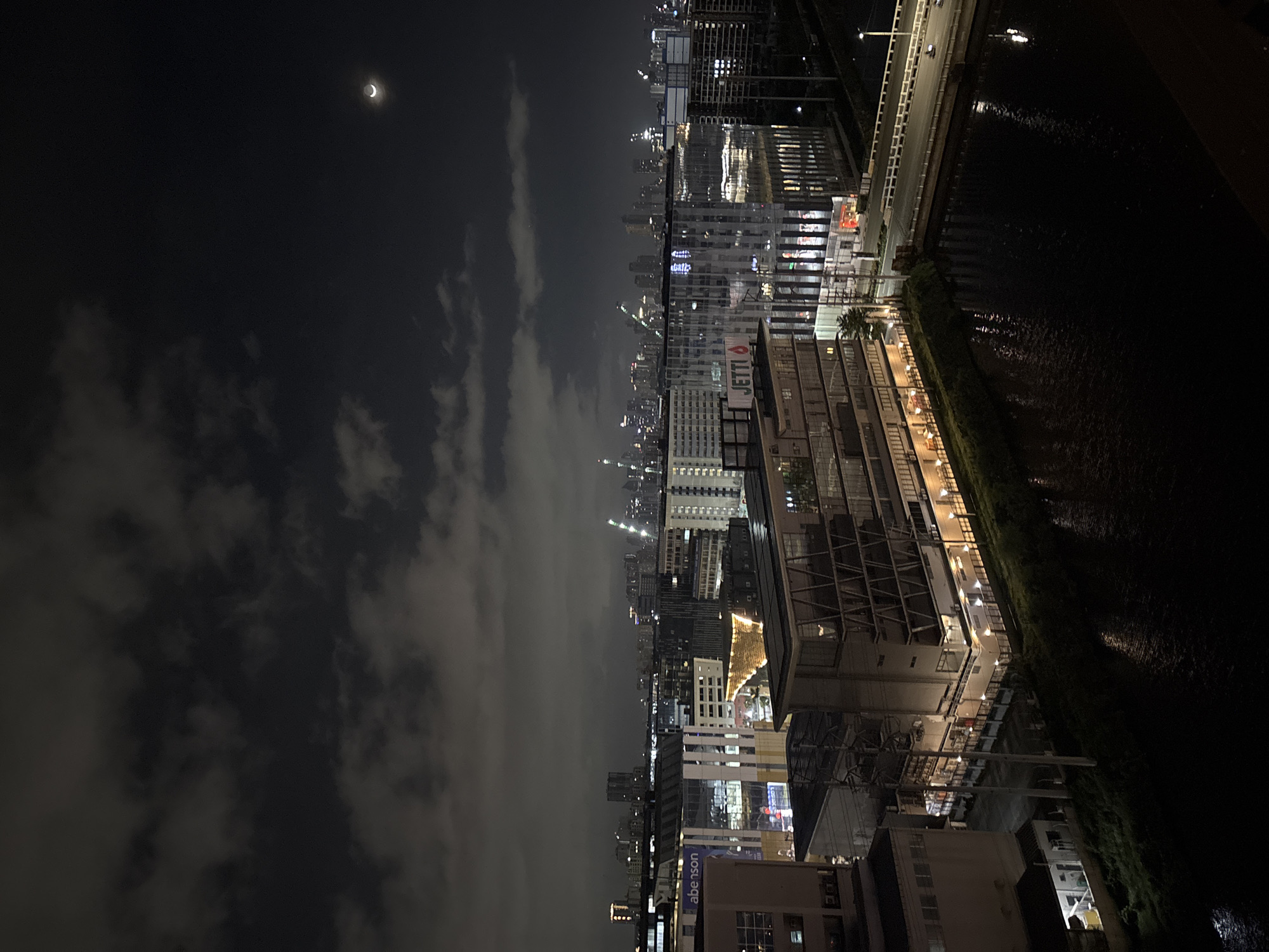 An aerial nighttime view captures a cityscape in Pasay, Philippines, characterized by illuminated buildings and a crescent moon in the cloudy sky. The scene is dominated by modern architecture, with various structures showcasing multiple stories and lit windows. Prominent buildings include a structure with a large "Jetti" sign and another with "Abenson" signage. The foreground features a dark body of water, likely a river or canal, reflecting the ambient light. Adjacent to the water is a green embankment lined with trees. A bridge crosses the water in the midground, with visible traffic. In the distance, a sprawling metropolis is visible with numerous high-rise buildings and construction cranes dotting the skyline, indicating ongoing development. The sky is partly overcast with dark clouds, partially obscuring the faint light of the crescent moon.