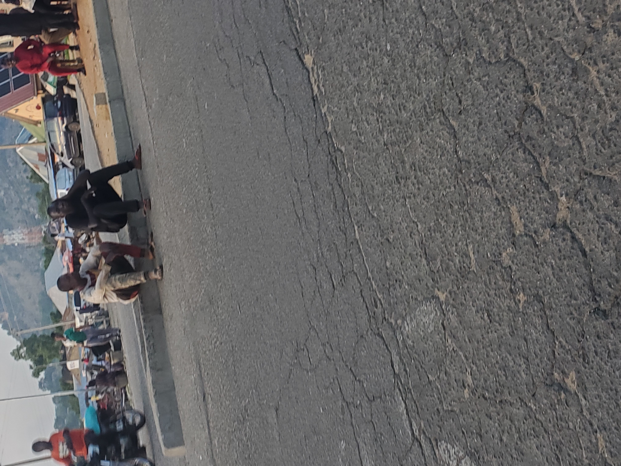 The image shows a street scene in Ampan, Nigeria. Several people are gathered on the side of a paved road, some sitting on the curb and others standing. In the foreground, a person in an orange shirt is seen next to a motorcycle. Further down the road, a group of people are clustered together, with some appearing to be engaged in conversation. The road surface is cracked and worn, suggesting it is a well-trafficked area. The background shows buildings and a hilly landscape, hinting at a semi-urban or rural setting. The lighting suggests it is daytime, possibly late afternoon, given the shadows. The overall atmosphere appears to be casual, with people appearing relaxed in their surroundings. No visible text is present in the image.