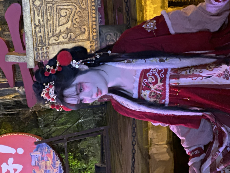 This image captures a young woman in elaborate traditional Chinese attire, standing in an outdoor, culturally themed environment in Yuzhong District, China, under ambient artificial lighting. The woman's dark hair is styled in an intricate updo, adorned with a central red and gold tiara-like piece, multiple red spherical ornaments resembling pom-poms, and dangling gold and silver hair ornaments featuring chains and small coin-shaped charms. Her face is made up with prominent pink eyeshadow, glitter accents on her cheeks, and pink lipstick. She wears a multi-layered outfit consisting of a red undergarment or dress with white and gold embroidered floral and bird motifs and gold tassels, covered by an outer red jacket trimmed with white fur, which exposes beige long sleeves underneath. She is positioned facing slightly right from the viewer's perspective, with a subtle smile. The background features a large red circular sign on the left with white Chinese characters and an illustration of an urban landscape. Behind and to the left of the woman, a textured green surface, possibly foliage or a rock face with water, is visible. To the right, a tall, ornate golden structure with embossed patterns stands. A dark metal chain runs horizontally across the lower mid-ground.
