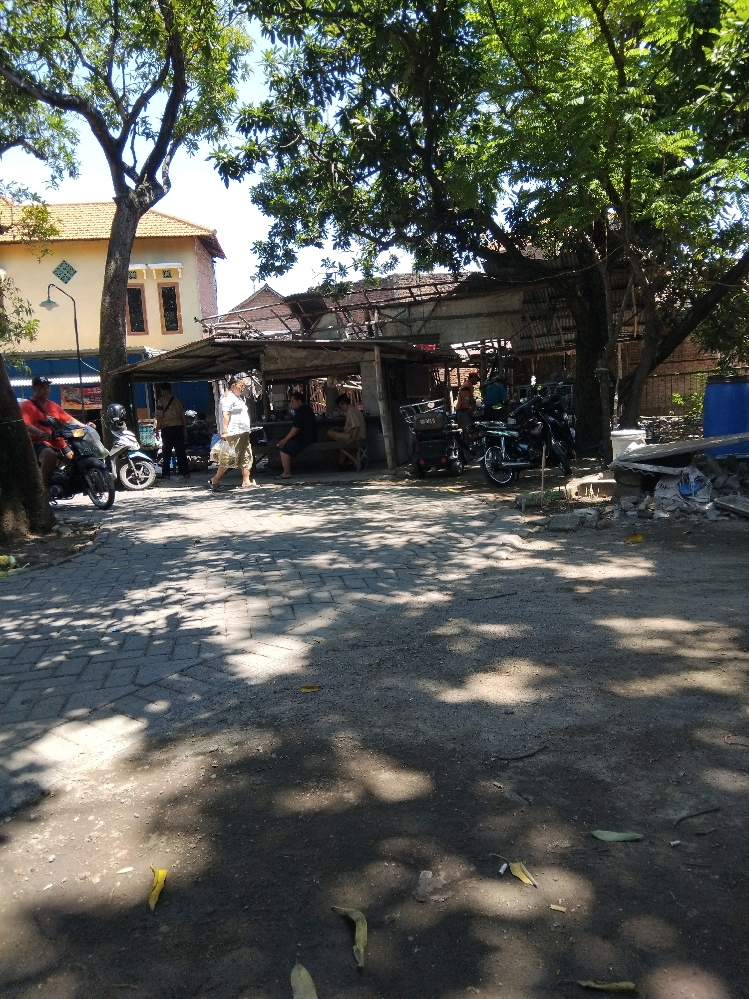 The image captures an outdoor scene in Kepuh Kiriman, Indonesia, under bright daylight, indicated by distinct shadows cast by trees onto the ground. The foreground is a dirt and gravel path, partially covered by paving stones. In the midground, a makeshift shelter with a corrugated iron roof is visible, supported by wooden structures. Several motorcycles are parked around this shelter, some of which appear to be for sale or repair.
Several individuals are present. To the left, a man in a red shirt is seated on a motorcycle, while another person stands nearby. Further into the shelter, a man in a white shirt and shorts is walking, carrying a plastic bag. Two other men are seated under the shelter, one in a dark shirt and another in a light-colored shirt, appearing to be engaged in conversation or activity. To the right of the shelter, a large blue water tank is situated, next to a pile of debris that includes broken roof tiles and wood. The background features a building with a yellow facade and orange roof tiles, with trees providing significant canopy cover throughout the scene. The overall environment suggests a casual, possibly commercial or residential area, given the presence of housing, vehicles, and a shaded gathering space.