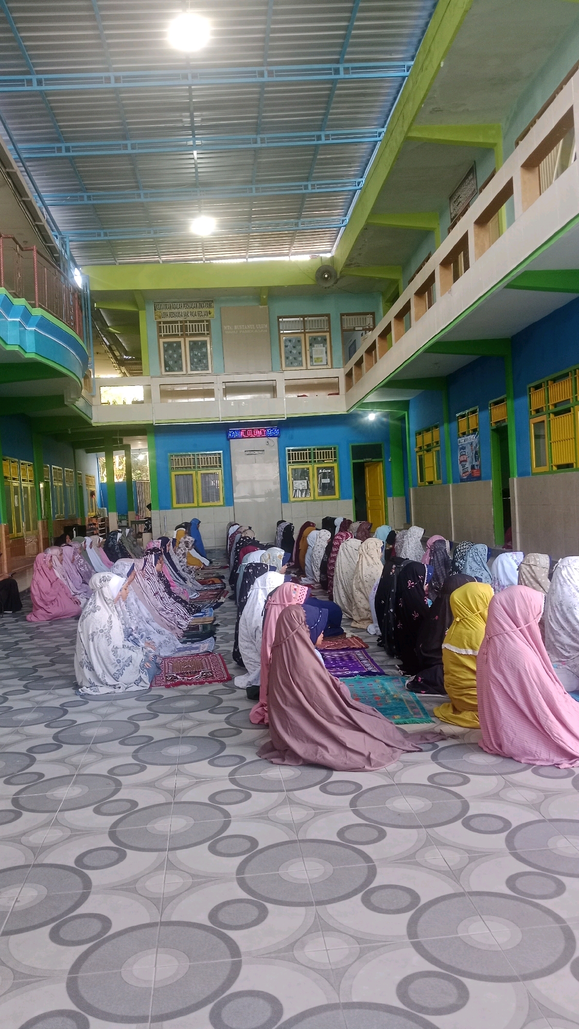 The image depicts an indoor scene, likely a prayer hall or school building, in Waru, Indonesia. A group of women, dressed in hijabs and modest attire, are engaged in prayer on individual prayer mats spread across a tiled floor. The floor is covered with a repeating pattern of concentric circles in shades of gray and white. The walls of the room are painted in bright colors, with sections of blue and green visible. Several windows, some with yellow frames, punctuate the walls. A blue-framed ceiling with exposed metal beams and fluorescent lights is overhead. In the background, signs with Indonesian writing are mounted on the walls, one of which reads "MTs. Bustanul Ulum Waru". The lighting is bright, suggesting daytime. The overall atmosphere is one of communal prayer and religious observance.