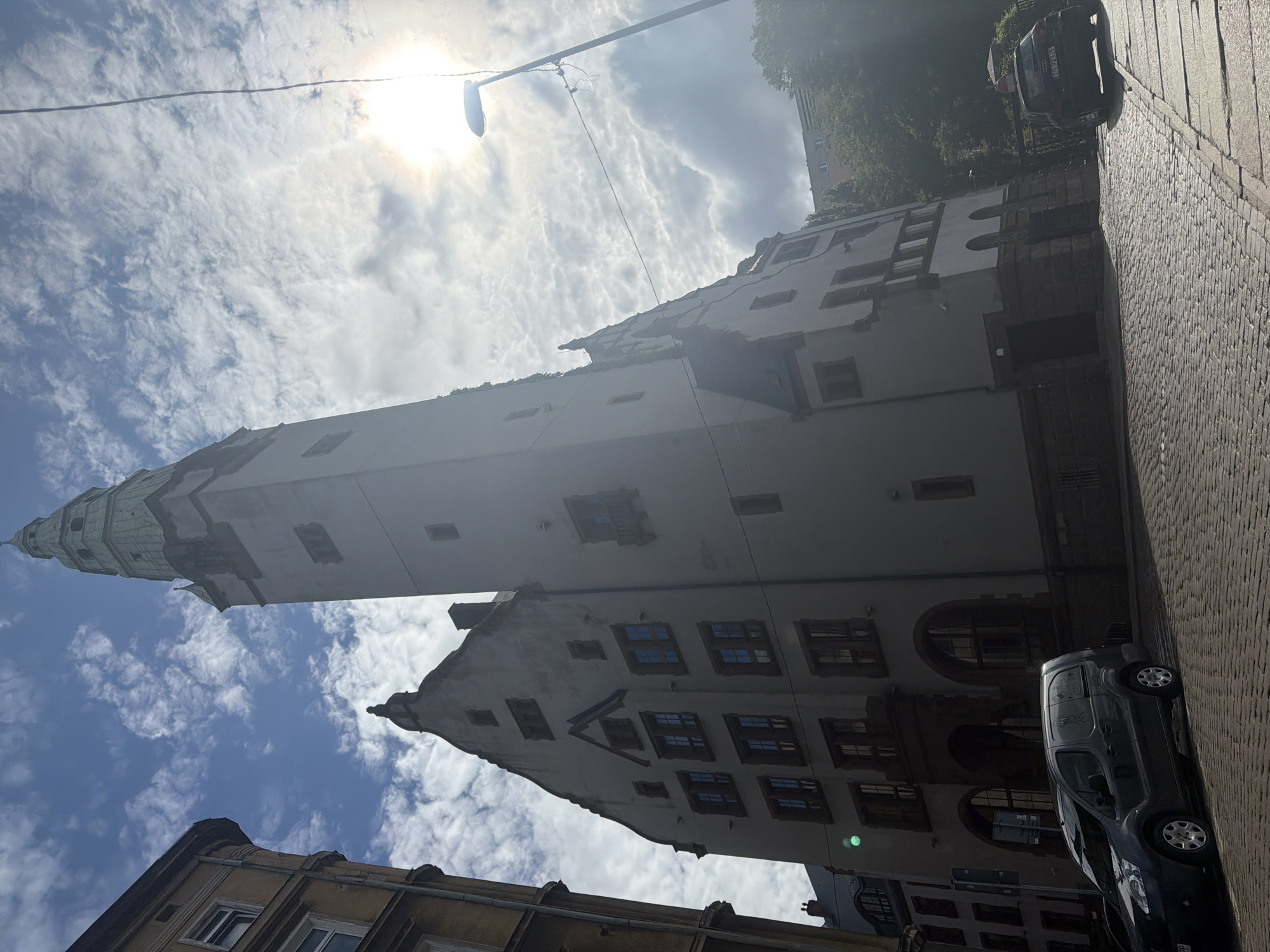 The image shows a street scene in Szczecin, Poland, featuring a large, imposing building with a tall tower. The architecture suggests a historical or institutional purpose, possibly a government building or a university. The building is made of light-colored stone or stucco and has a green-tiled spire atop its tower. The facade is adorned with numerous windows and decorative architectural elements.
In the foreground, a cobblestone street is visible, with a dark-colored van parked on the left side and a dark-colored car on the right. The van appears to be a commercial vehicle, possibly for deliveries or services. The car on the right has a license plate that is partially visible.
The sky is partly cloudy with the sun shining brightly, creating some glare and lens flare. A street lamp is also visible on the right side of the image. Trees and other buildings can be seen in the background, indicating an urban environment. The overall impression is of a sunny day in a city with a mix of historic and modern architecture.
The building in the center of the image is likely the Pomeranian Dukes' Castle (Zamek Książąt Pomorskich) in Szczecin. This landmark is characterized by its tall tower and historical architecture, matching the visual elements in the photograph.