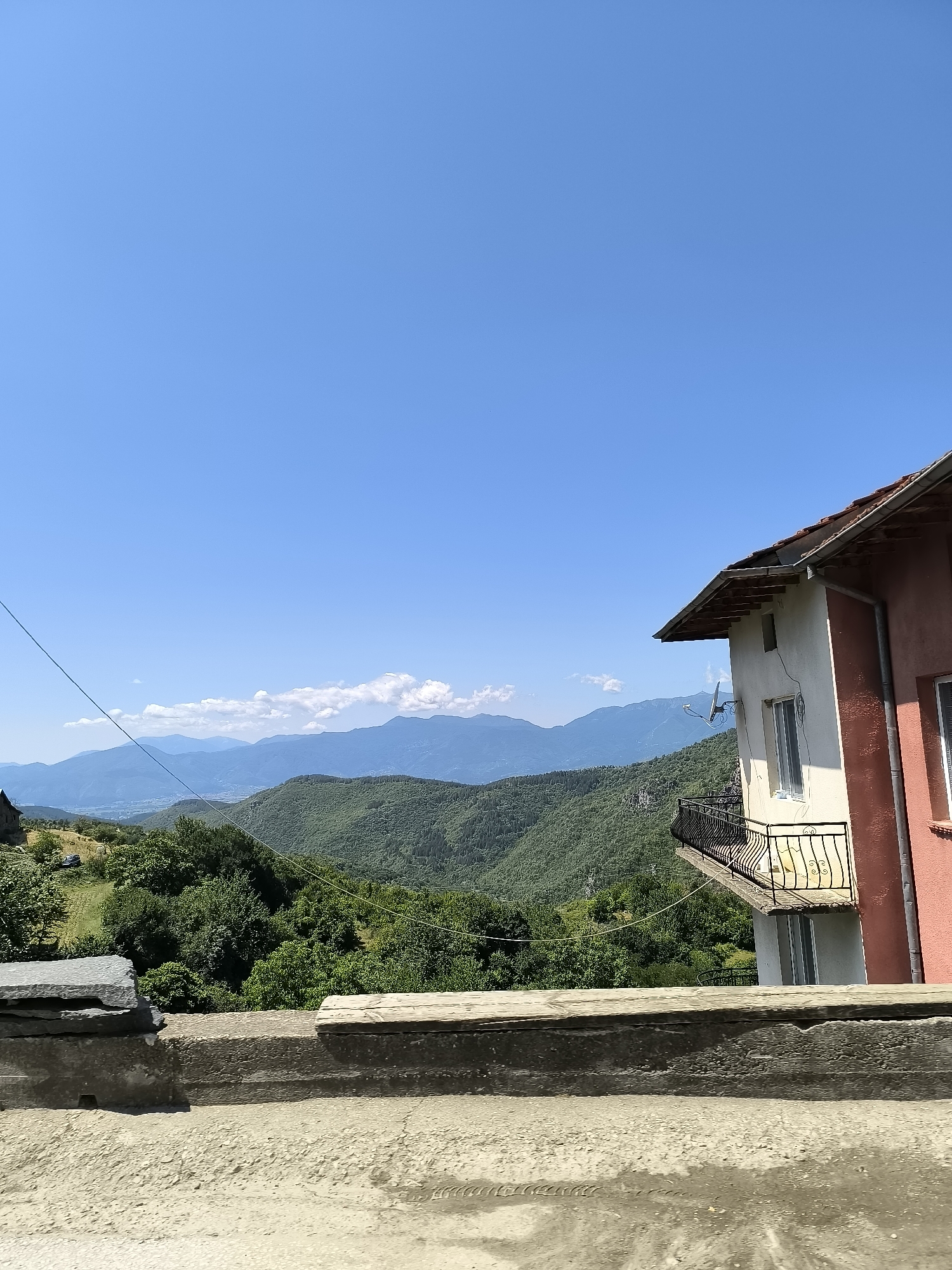 The image shows a scenic view of the village of Leshten, Bulgaria, on a clear day. In the foreground, there is a concrete structure with a stone and a wooden plank. To the right, a portion of a two-story house is visible, featuring a pink and white facade, a red tiled roof, a balcony with a black metal railing, and satellite dishes. The background is dominated by rolling green mountains covered in lush forests, with layers of mountains receding into the distance under a bright blue sky. A few white clouds are scattered across the sky. A thin wire, likely a utility line, stretches diagonally across the frame. The overall impression is one of a tranquil, rural mountainous landscape.