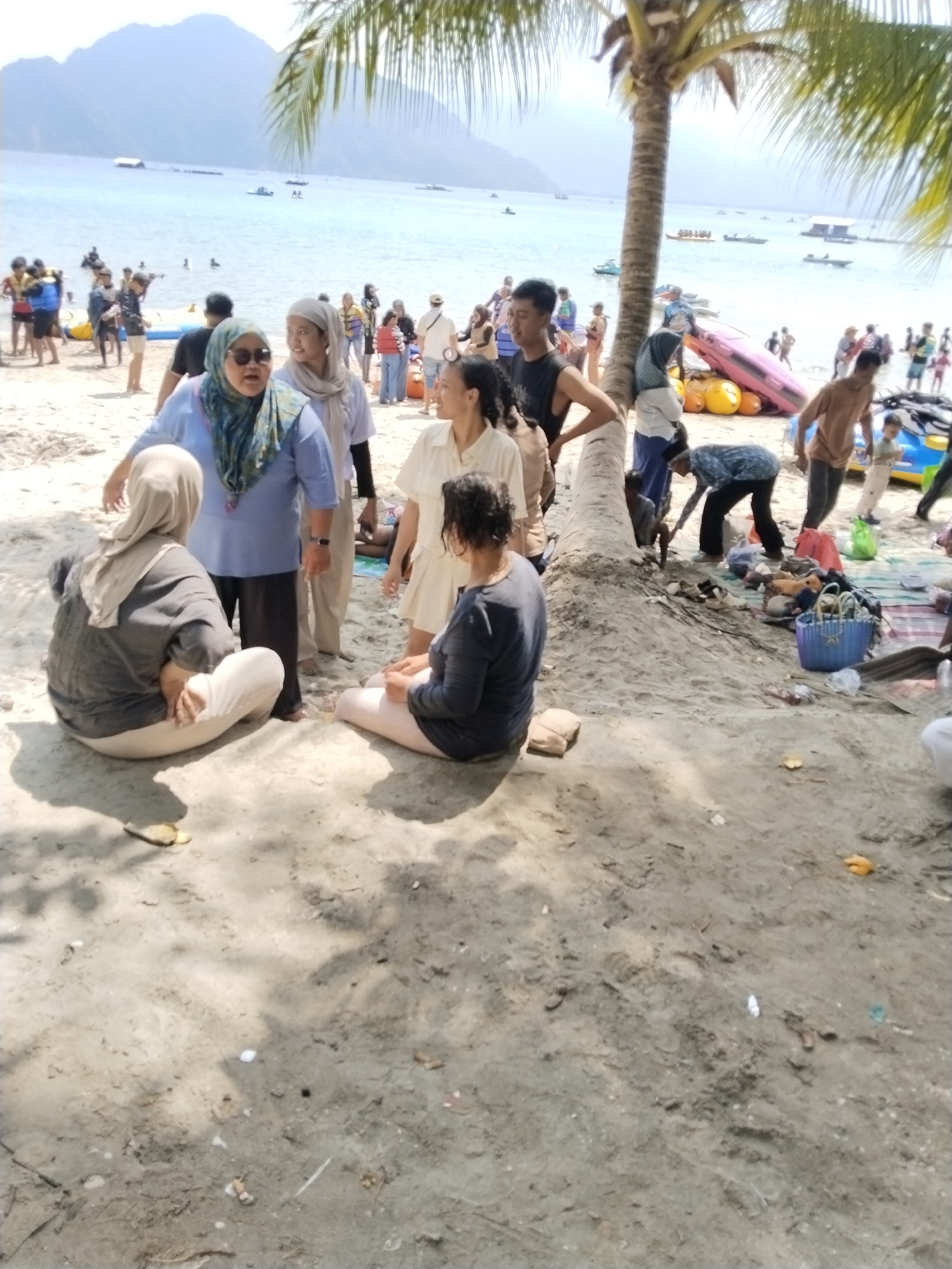 This image depicts a busy beach scene in Prigi, Indonesia, with numerous people enjoying a sunny day. In the foreground, several individuals are gathered on the sand. One woman, wearing a grey headscarf and a dark long-sleeved shirt, is seated with her legs crossed, facing away from the camera. Another person, also seated, is wearing a dark long-sleeved shirt and light-colored pants. Standing nearby is a group of people. A woman in a blue headscarf and sunglasses is talking to another woman in a light grey headscarf. Behind them, a man in a black sleeveless shirt and shorts is visible.
Further in the background, the beach is populated with many more individuals. Some are wading in the clear blue water, while others are on the sand near boats and inflatable watercraft. The water is calm with several boats on the horizon. Mountains rise in the distance, providing a scenic backdrop. A prominent palm tree with its fronds casting shadows on the sand is situated in the middle ground. The overall atmosphere suggests a lively tourist destination.
