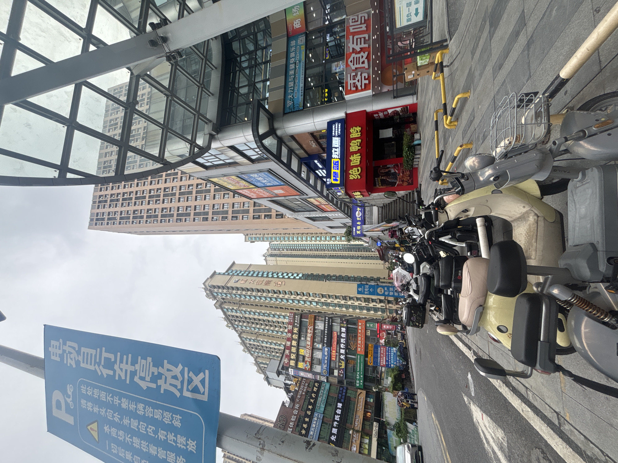 The image depicts an outdoor scene on a street in Nanhai District, China. In the foreground, several parked electric scooters and motorcycles line the sidewalk. A blue and white parking sign with Chinese characters indicating a "Electric Bicycle Parking Area" is visible on a pole to the left. The street is paved with grey and white striped lines, and a yellow barrier is placed along the edge of the sidewalk. On the right side of the image, a modern building with a glass facade and a metal frame is present. Multiple stores with illuminated signs in Chinese characters are situated on the ground floor of the building. In the background, tall residential buildings with numerous windows rise into the sky under an overcast sky. A few people are faintly visible on the street and sidewalk, and a car is parked on the left side of the road. The overall impression is a bustling urban environment with commercial and residential areas.