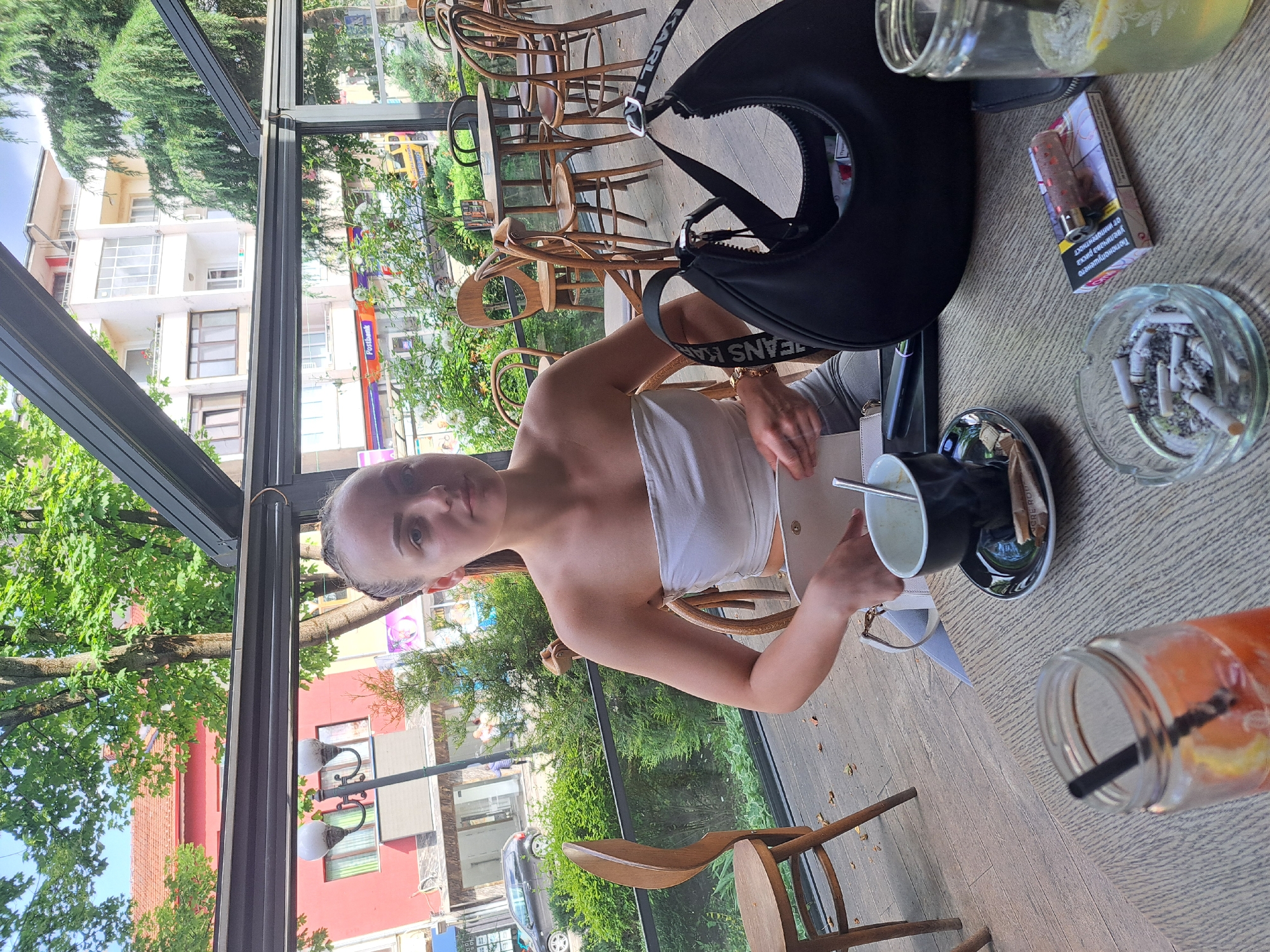 A person is sitting at a table in an outdoor cafe in Troyan, Bulgaria. The person is fair-skinned with light hair. They are wearing a white top and holding a small, light-colored purse. On the table are a black bag, a cigarette pack, an ashtray filled with cigarette butts, two drinks in glass jars, and a cup with a saucer. Wooden chairs surround the table. The cafe is enclosed by a glass structure, and outside there are trees, buildings, and a street with cars.