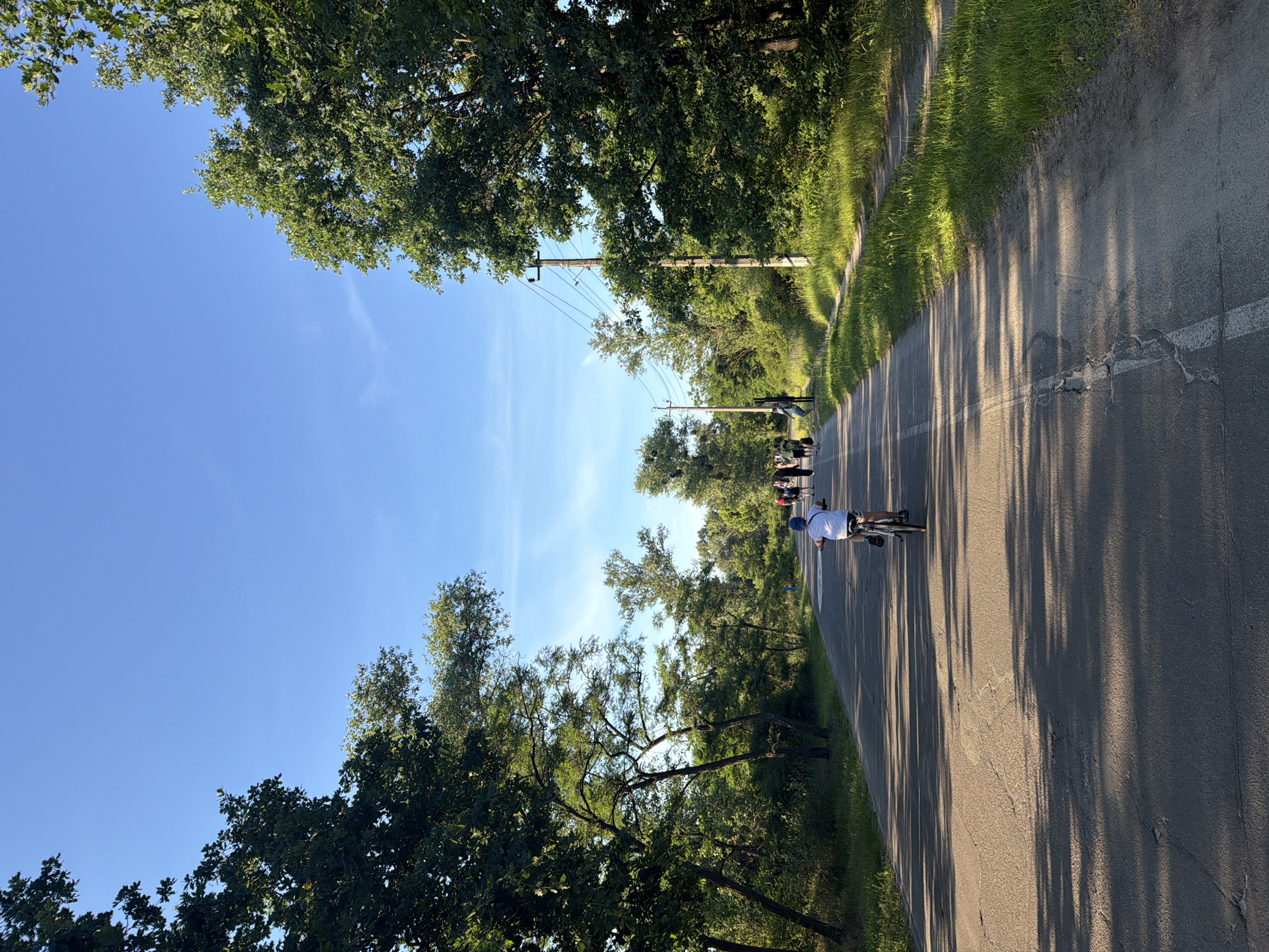 This is a photograph taken from behind a person riding a bicycle on a paved road in Kyiv, Ukraine. The road is lined with trees on both sides, with lush green foliage. The sky is clear and blue, with a few wispy clouds. In the distance, a group of people can be seen walking on the side of the road. There are also power lines visible, supported by wooden poles. The lighting suggests it is late afternoon or early evening, with long shadows cast across the road by the trees. The overall impression is of a peaceful outdoor scene in a park-like setting within the city.