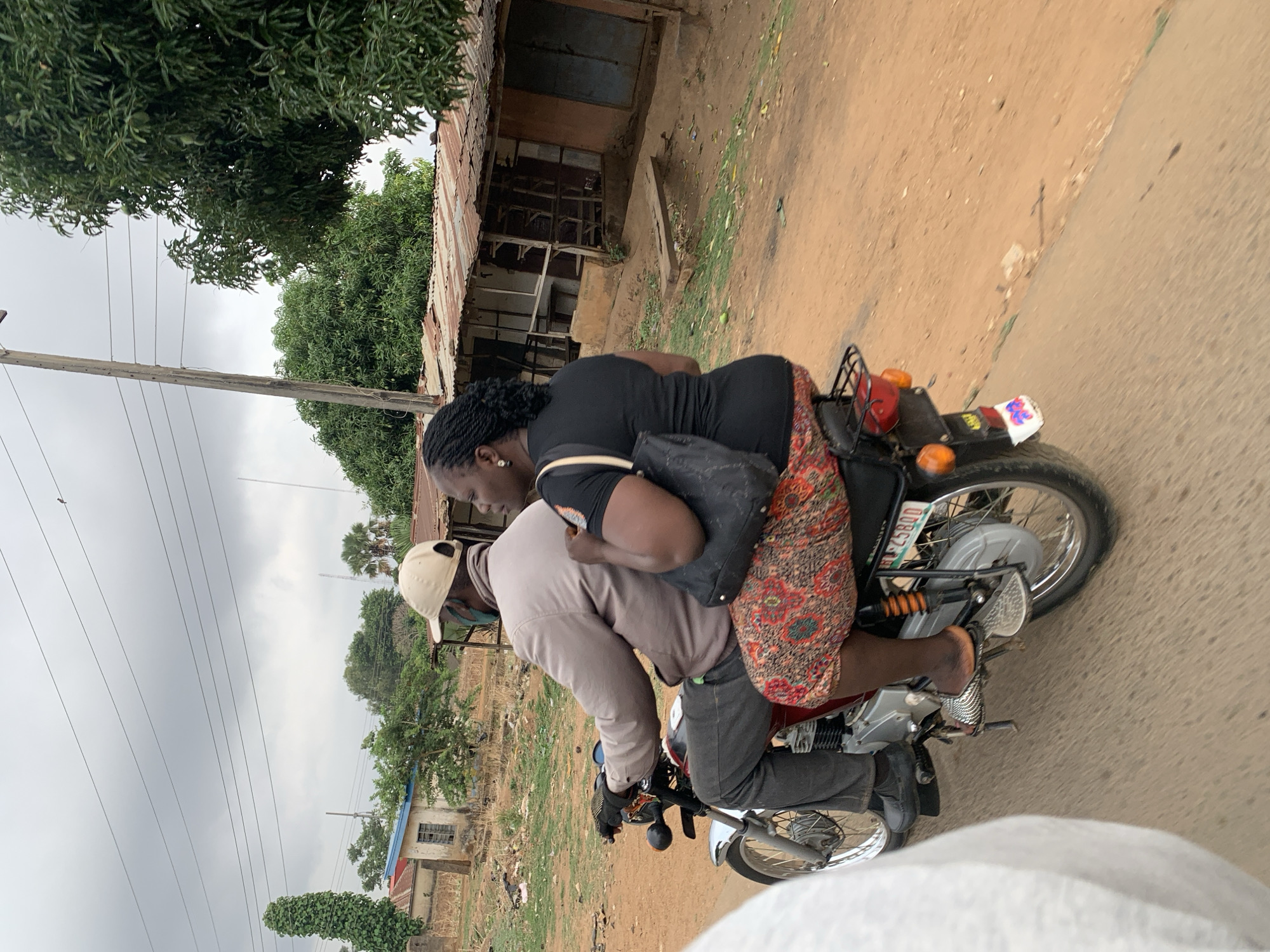 Two individuals are riding a motorcycle on a dirt road in the city of Maigizo, Nigeria. The driver wears a light brown jacket, black gloves, dark pants, and a tan baseball cap. The passenger, seated behind the driver, wears a black top, a patterned skirt, and carries a black bag. The motorcycle is red with silver accents. The background shows foliage, utility poles, and a dilapidated building. The sky is overcast.