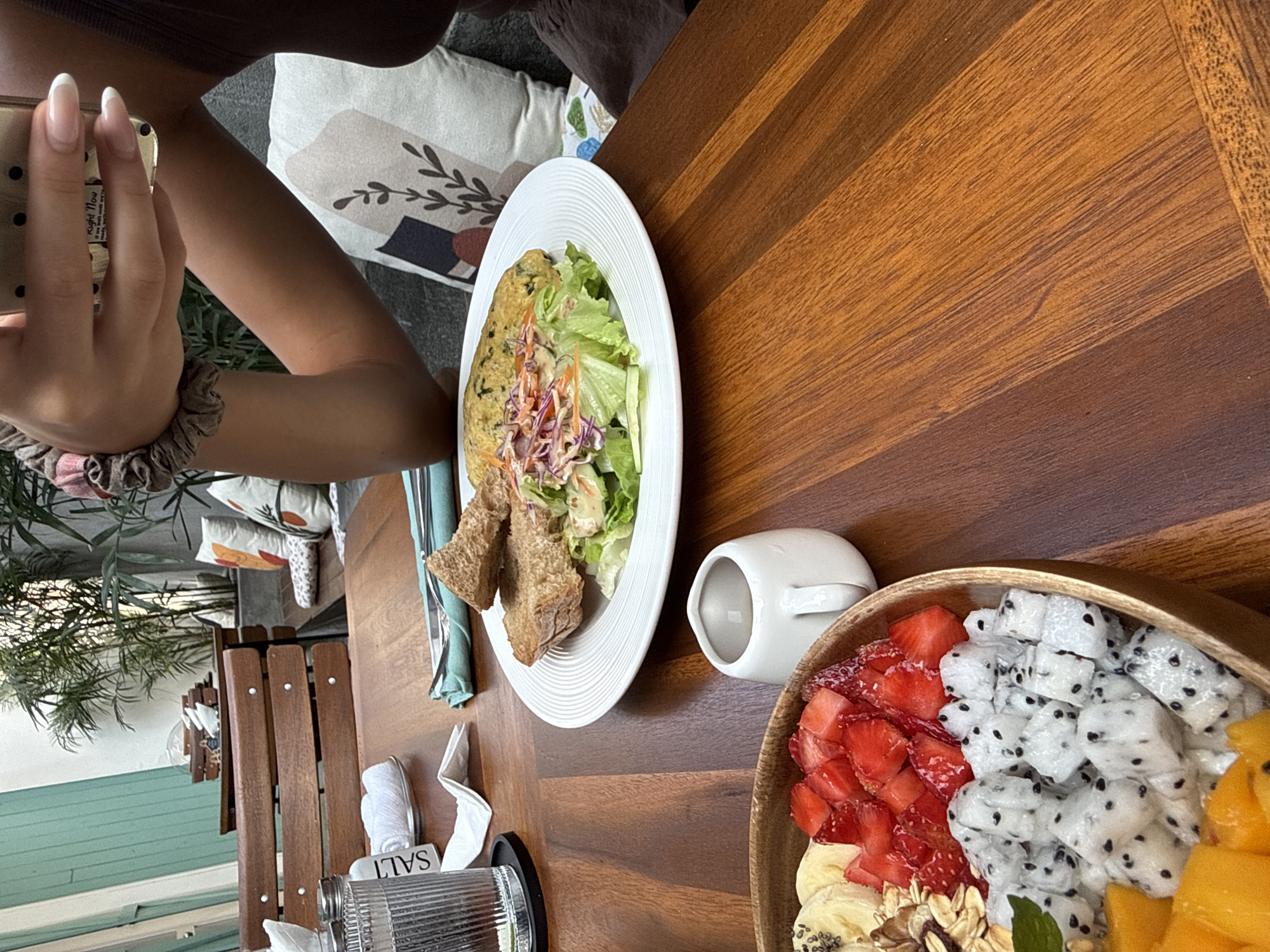 The image shows a person seated at a wooden table, likely in a restaurant or cafe, taking a photo with a phone. The table has a plate of salad with bread, a small white creamer, and a wooden bowl of fresh fruit including strawberries, dragon fruit, mango, and banana. Utensils and condiments are also visible on the table. The location may be Đà Nẵng, Vietnam based on common elements and the presence of tropical foliage.