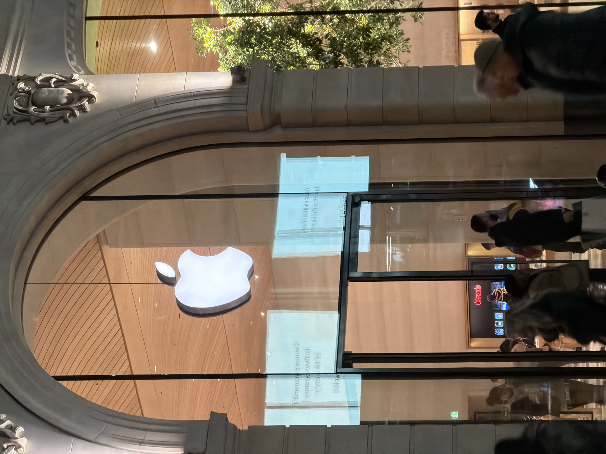 Captured from an exterior vantage point at night, the image showcases an Apple Store nestled in London, United Kingdom. The store front, characterized by a prominent archway adorned with intricate moldings, features a large glass panel displaying the illuminated Apple logo against a light-colored wooden backdrop. The glass reflects passersby, including individuals in dark clothing, who appear blurred due to movement. Inside, screens display "Arcade" and other content. Above the archway, hints of verdant trees and building facade details suggest the store's urban setting. The scene blends architectural elegance with modern retail, capturing a moment in the city.