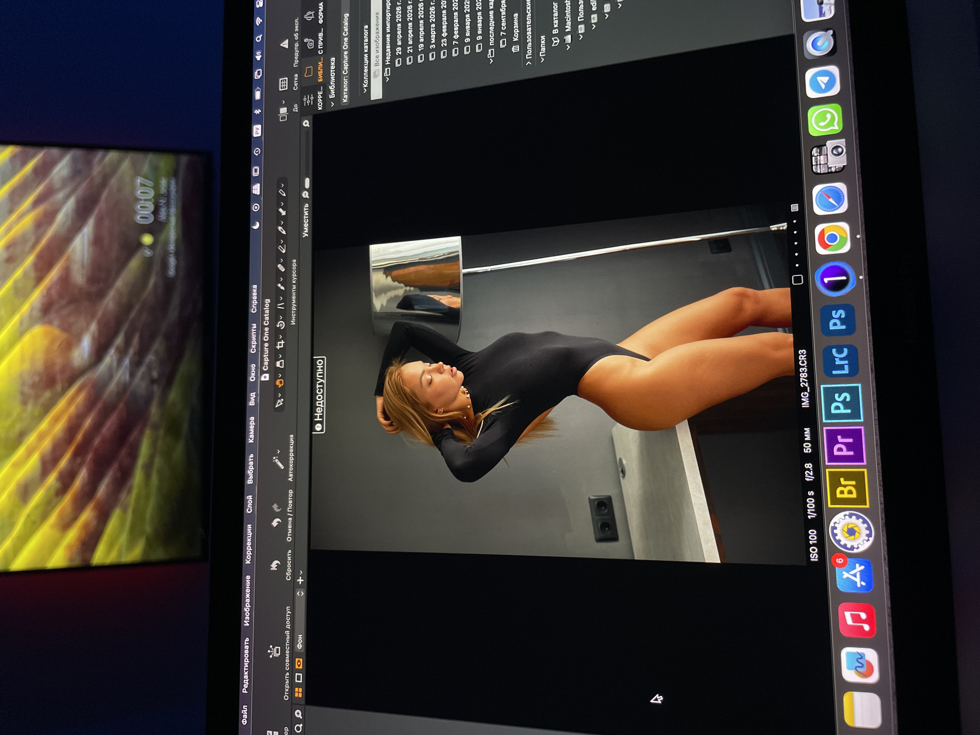 The image is a photograph of a computer screen, taken at an angle, displaying a software interface and other elements. The software appears to be a photo editing application like Adobe Lightroom or Capture One, showing an image of a woman lying down wearing a black bodysuit, and facing the camera. The display shows the image editing interface, with various adjustment tools and image metadata visible along the right edge. The screen also has a list of catalog entries on the left side. The bottom edge of the screen shows various applications docked in the tray. The computer screen's background wallpaper shows 00:07, and is a bright yellow design. The photograph appears to have been taken indoors, and a portion of another display is visible in the lower left of the frame, possibly reflecting a colorful abstract scene. There is no evidence to suggest any location or city.