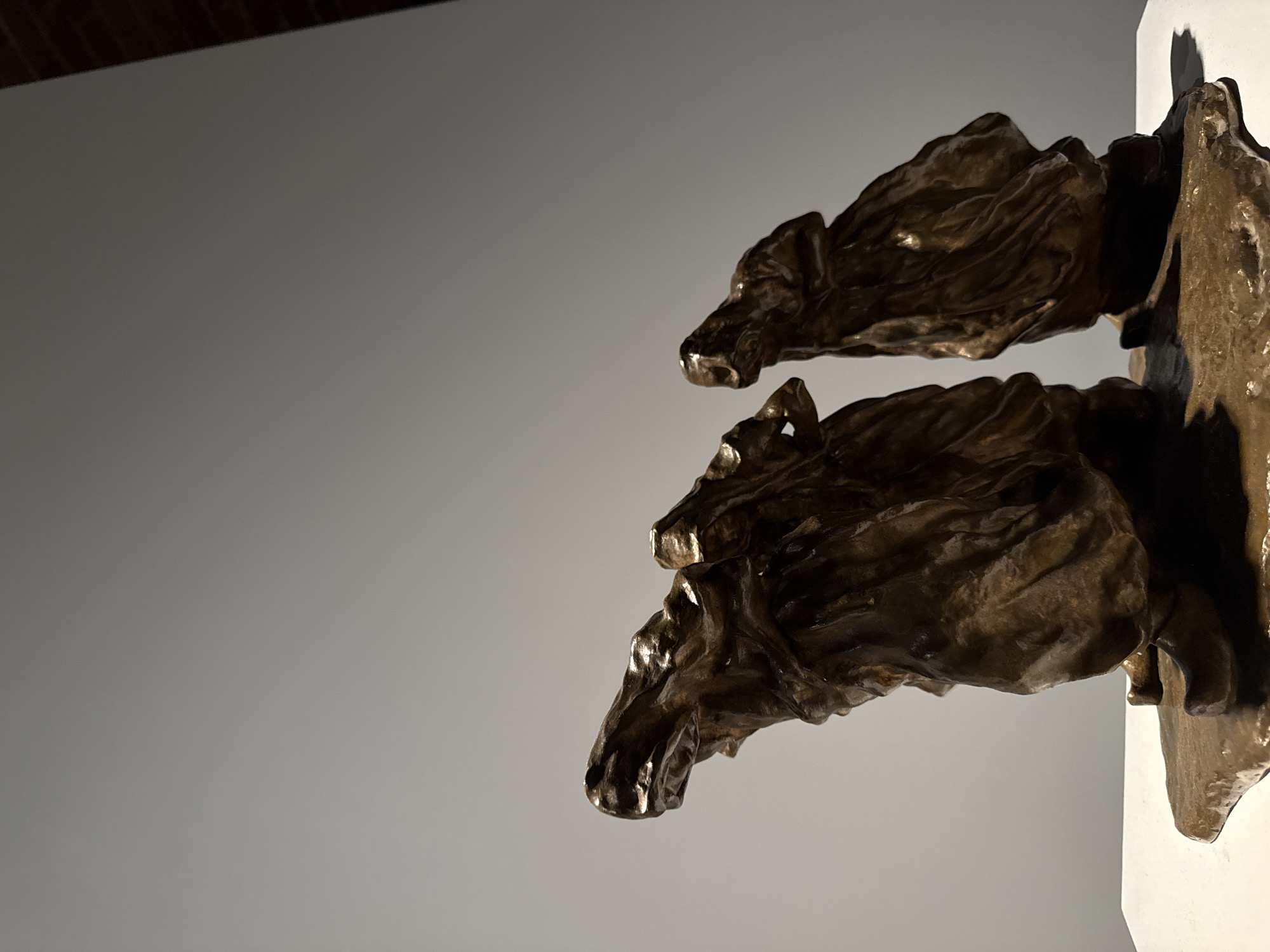The image shows a bronze sculpture of two bear heads emerging from a white wall or support. The sculpture is located in Nizhny Novgorod, Russia. The bronze has a textured, almost wrinkled appearance, and it is shiny in places due to the lighting. The background is a plain gray wall, and the angle of the photo is slightly tilted.