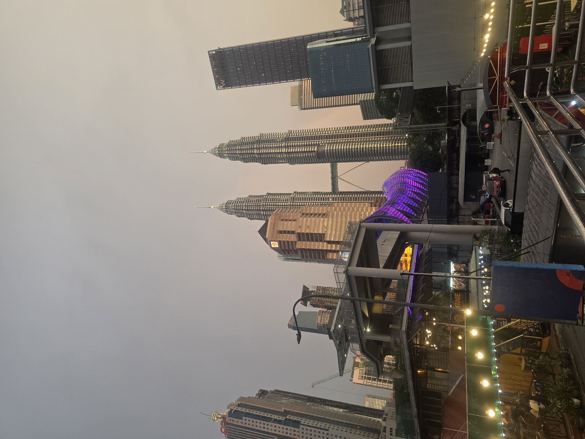 The image displays a bustling urban cityscape in Kuala Lumpur, Malaysia, under a hazy, overcast sky. Dominating the mid-ground are the iconic Petronas Twin Towers, their intricate silver structures rising prominently above surrounding buildings. Nearby, several other modern skyscrapers contribute to the dense urban fabric, with one tall, dark building featuring the letters "RW" visible near its summit.
An elevated pedestrian bridge or walkway spans across the mid-ground, distinguished by a segment brightly illuminated with vibrant purple lights, reminiscent of the Saloma Link Bridge. Below this elevated structure, the street level features buildings and establishments, likely restaurants or cafes, with outdoor seating areas. These areas are warmly lit by numerous string lights, creating an inviting ambiance.
The sky is a muted, desaturated orange-grey, suggesting either significant atmospheric haze, smog, or the specific light conditions of dusk or early evening. The artificial lights from buildings, streetlamps, and the decorative string lights are all active, indicating a transition from day to night. Visible text includes "RW" on a skyscraper, "BIRYANI" on a street-level sign, and "KL" on another sign. Although individual people are not distinctly visible, their presence is implied by the active street scene and outdoor dining areas. The overall impression is a vibrant metropolis at twilight, characterized by its architectural landmarks and lively street life despite the hazy conditions.