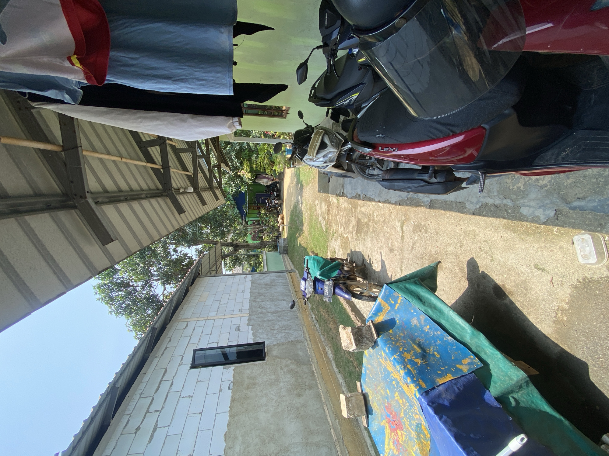 The image depicts an outdoor scene in Batam, Indonesia, likely a residential area or a small business frontage. In the foreground, a row of parked motorcycles is visible. The closest motorcycle is partially obscured, but its red bodywork and black seat are discernible. Behind it, a black and silver motorcycle is parked, followed by a dark blue motorcycle with a green cover on the seat and a license plate that reads "BP 2308 H". To the left of the motorcycles, a table covered with a blue and yellow tarp, supported by green tarps, is set up. A blue and white brick building is visible on the left side of the frame, with a single, narrow, dark-framed window. The building's lower section appears to be plastered or stuccoed. A corrugated metal roof extends over the motorcycles and the building, supported by metal beams and wooden poles. Clothes, including a light blue garment with red trim, a white garment, and a black garment, are hanging on a line under the roof. The ground is paved with concrete and some patches of grass are visible. In the background, more trees and a partially visible green building can be seen. The sky is clear and blue, suggesting a sunny day. The overall impression is of a functional, everyday outdoor space.
