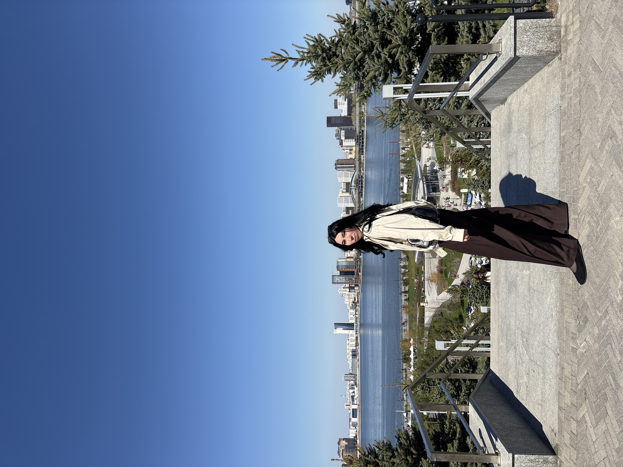 A young woman is the primary subject of the image. She is standing on a set of stairs with a cityscape and a body of water in the background. The woman is wearing a light-colored jacket and a long, dark skirt. She has dark hair and is looking directly at the camera. The setting is outdoor, and the clear blue sky suggests a sunny day. The foreground features a paved area, likely a pedestrian walkway or plaza. A metal railing runs along the edge of the walkway, overlooking the scene below. Below, there is a river with a park and modern buildings on either side. The cityscape features a mix of high-rise structures, indicating an urban environment. The city is Kazan, Russia.