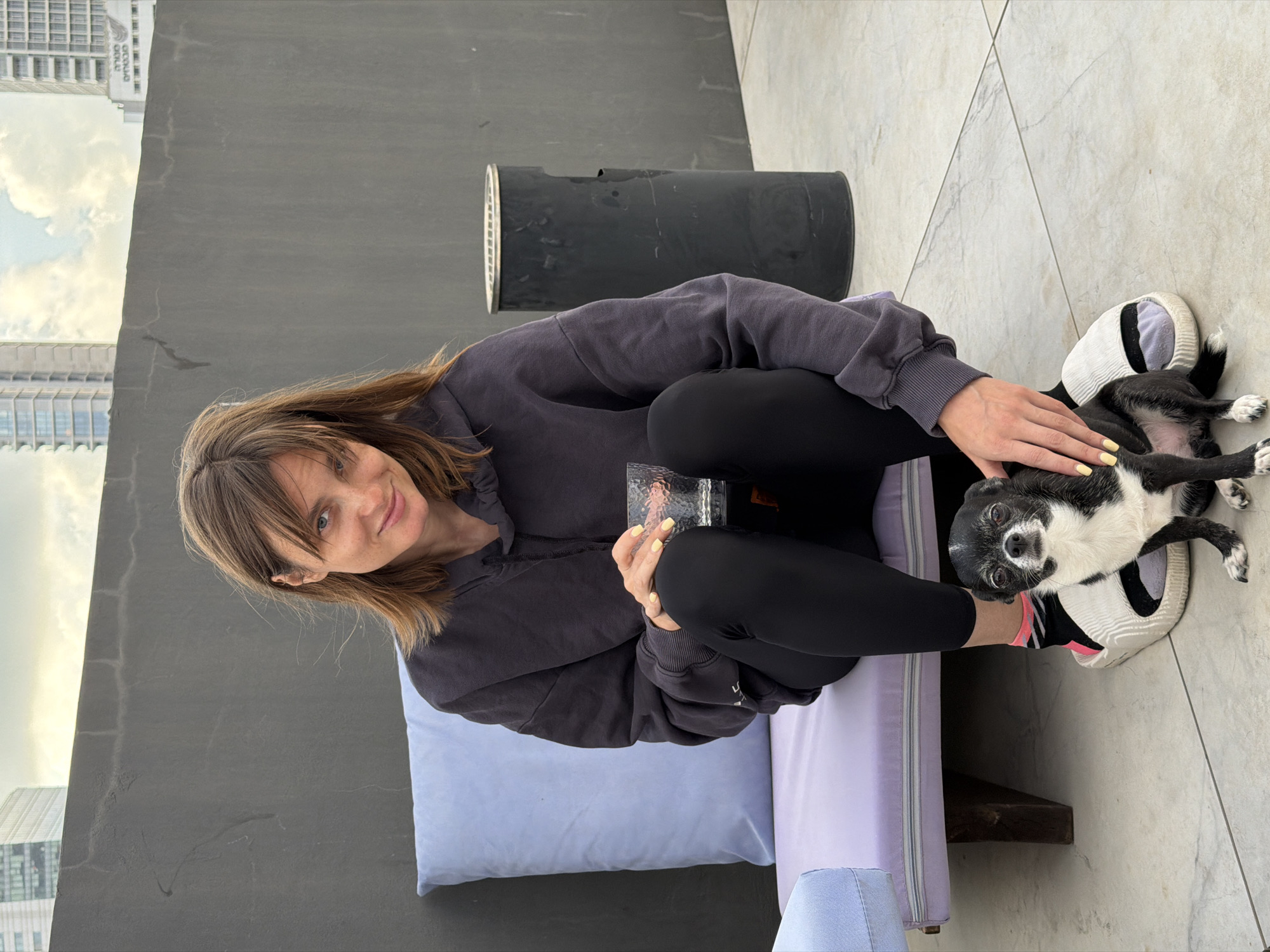 A woman is sitting cross-legged on a lavender-colored, outdoor bench on a rooftop terrace in Tel Aviv, Israel. She is wearing a dark grey hooded sweatshirt and black leggings. In her hands, she holds a clear glass. A black and white dog lies on its back near the woman's feet, which are propped on a pair of white athletic shoes. The woman is petting the dog. To her left, a large, cylindrical, black object sits on the terrace. In the background, buildings typical of Tel Aviv's urban landscape are visible, with a cloudy sky above. The terrace surface is composed of light-colored tiles.