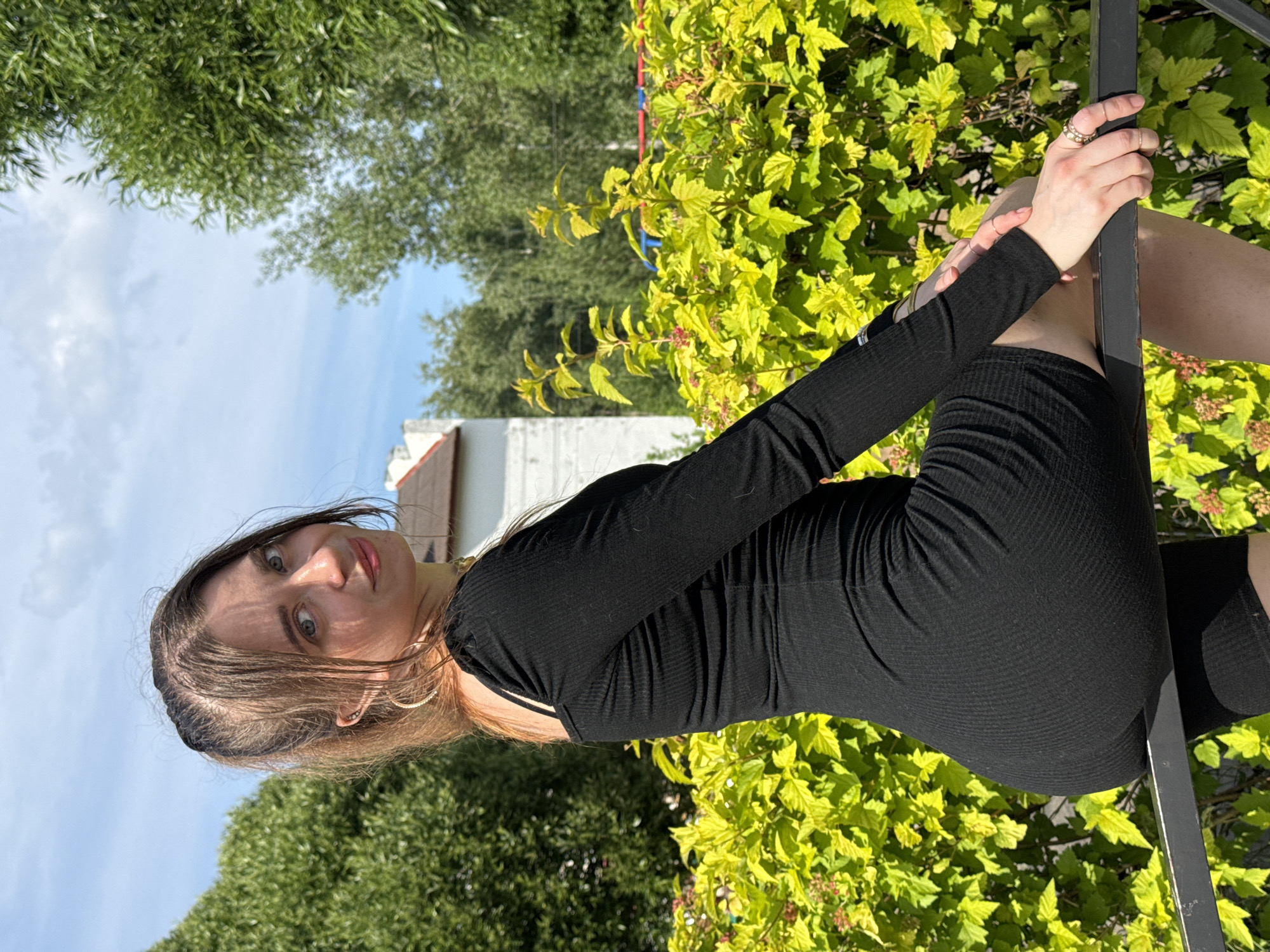 A young woman with brown hair, wearing a black ribbed long-sleeved dress, is posing outdoors. She is leaning against a dark metal railing, with a lush green bush behind her. The sun is shining, casting shadows and highlighting the vibrant colors of the foliage. Her hair is slightly windswept, and she has a thoughtful expression on her face. She is wearing rings on her fingers and hoop earrings. The background also shows a glimpse of a white building and trees under a clear blue sky, suggesting a daytime setting. The scene appears to be tranquil and relaxed, with the woman appearing comfortable and confident. The image is oriented vertically.