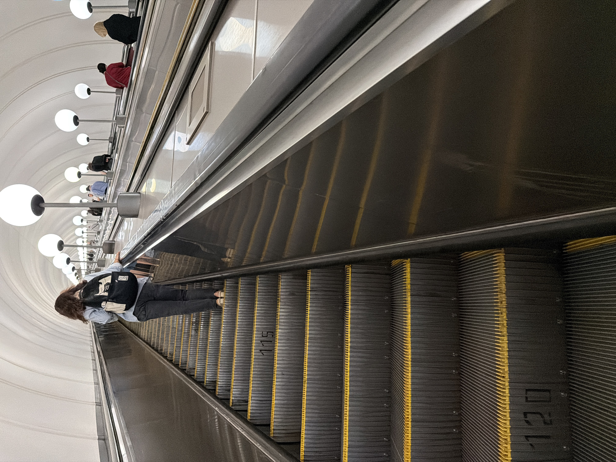 The image depicts a wide, well-lit escalator in a Moscow metro station, likely deep underground due to the absence of natural light and the tunnel-like architecture. The escalator is moving upwards, with several people ascending. In the foreground, a woman with dark, wavy hair is seen from behind, wearing a light blue shirt, dark pants, and a black and white backpack. She is walking up the escalator. Further up the escalator, other individuals are visible, some seated and others standing. The escalator steps are dark gray with bright yellow safety lines. The ceiling is a white, vaulted structure with rows of large, spherical white lights illuminating the space. The side of the escalator features a polished metal handrail and a metallic wall. The numbers "115" and "120" are stenciled on some of the escalator steps. The overall impression is one of a functional and well-maintained public transit environment.