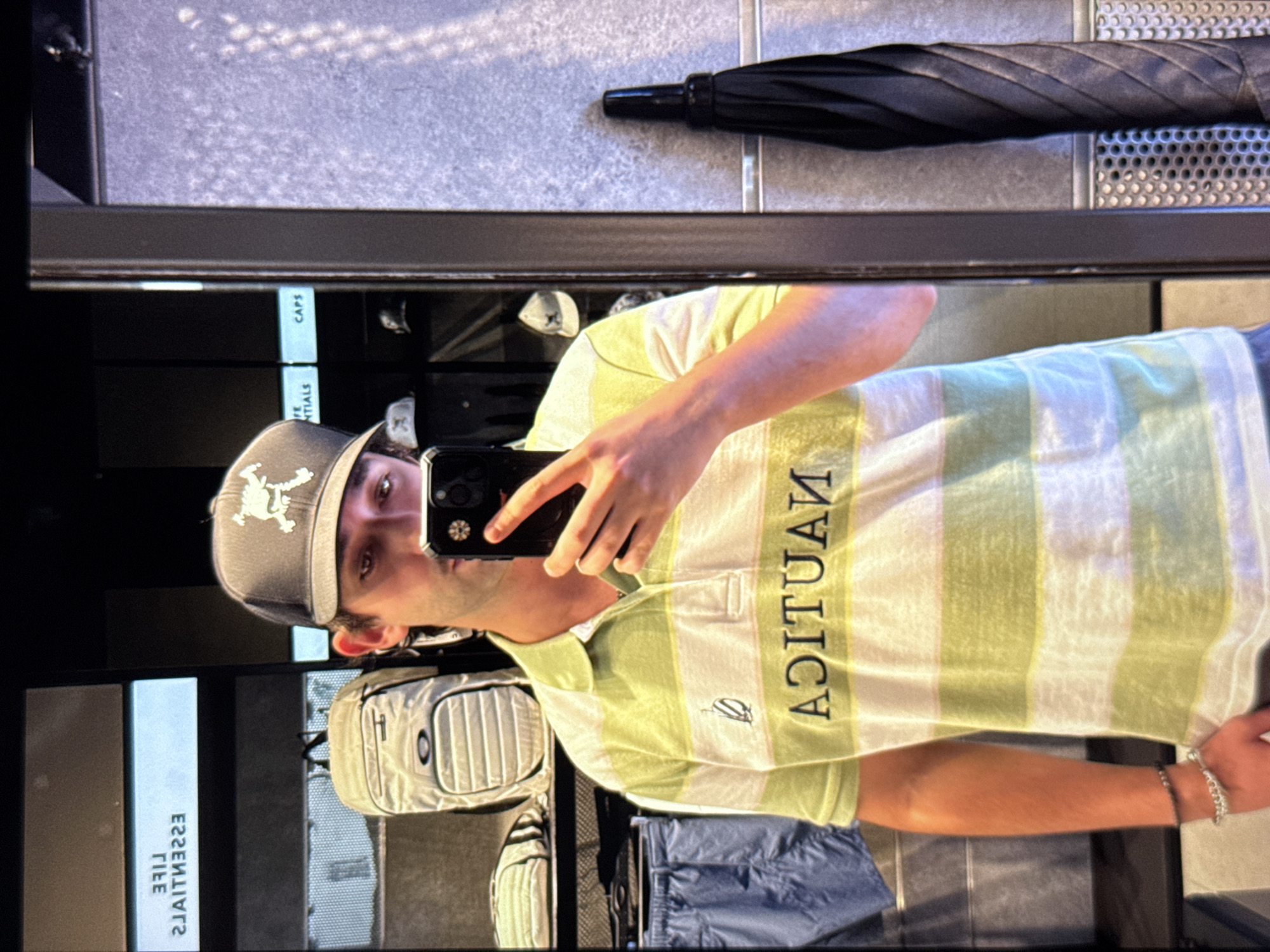 A person is standing in front of a mirror, taking a selfie. They are wearing a light green and white striped polo shirt with the Nautica logo on it and a gray baseball cap with a white skull logo. They have on two bracelets on their left wrist. In the background, shelves are visible with backpacks and hats on them, and what appears to be a black umbrella is leaning against a textured wall. The reflective surface of the mirror also shows a sign that reads "LIFE ESSENTIALS CAPS." The person's attire and the display of merchandise suggest they are in a retail store.