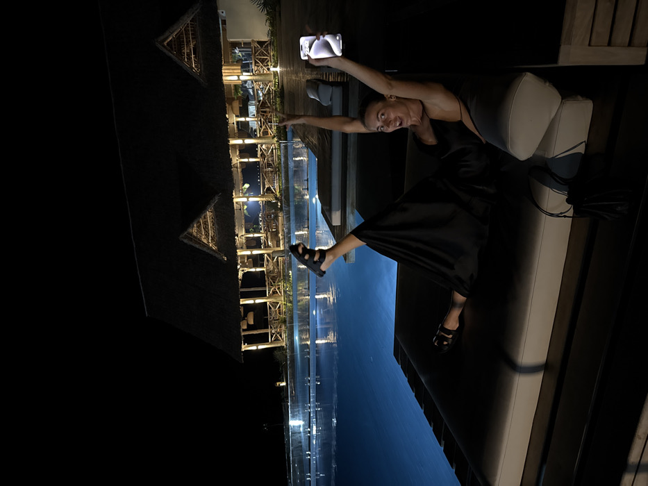 A woman with short hair poses for a nighttime selfie, holding a phone above her head. She wears a black dress and sandals, sitting on a cushioned outdoor lounge chair. Behind her is a dark, reflective pool of water reflecting the lights of a multi-story wooden structure with thatched roofs. The structure appears to be a building with multiple levels and illuminated openings. The background is primarily dark, with the lights from the building and pool illuminating the scene.