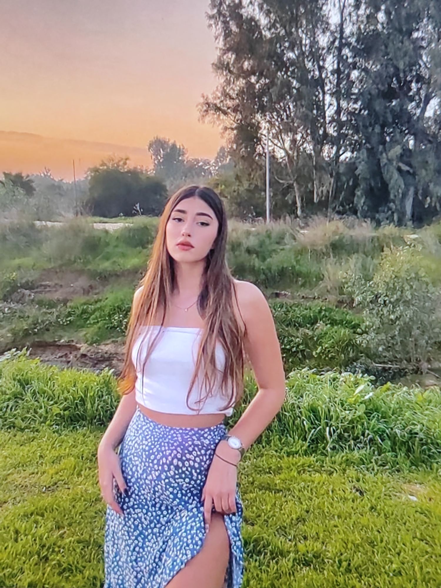 A young woman stands in a grassy field, wearing a white tube top and a blue skirt with a white floral pattern. Her long, brown hair cascades down her shoulders. She has a serene expression on her face and is looking directly at the camera. On her left wrist, she wears a watch.
The background is a natural landscape with trees and foliage, illuminated by the warm glow of a sunset. The sky displays shades of orange and yellow, suggesting it is dusk. The terrain appears slightly uneven, with patches of dry earth and lush green grass. The overall setting is outdoors and natural, possibly a park or a rural area.
The image is a medium shot, focusing primarily on the woman and her attire, with the natural environment serving as a backdrop. There is no visible text in the image. The mood is peaceful and natural, highlighted by the soft lighting of the sunset.