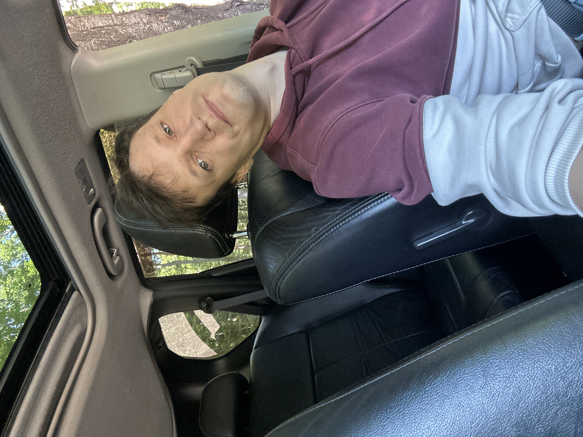 A man is taking a selfie in a car with the sunroof open. He is wearing a maroon hoodie with a cream-colored long-sleeved shirt underneath. The car's interior appears to be black leather. Trees and foliage are visible through the sunroof and windows, suggesting an outdoor setting, possibly in a wooded or rural area. The man has short brown hair and light-colored eyes, and he is looking directly at the camera with a slight smile. There are no other individuals or objects of interest clearly discernible in the image besides the car's interior and the man. The image does not contain any identifiable landmarks or features that would specifically place it in Shatava, Ukraine.