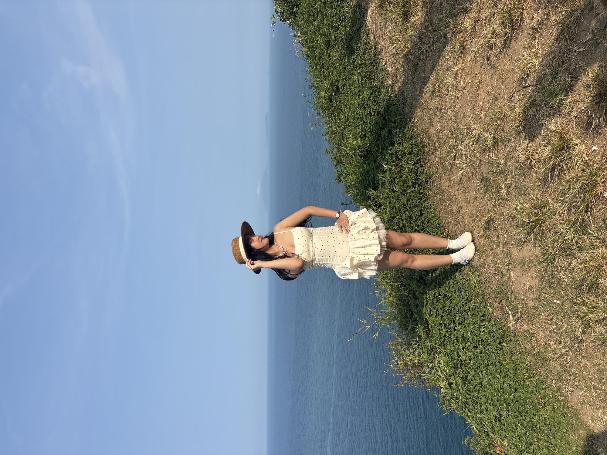A person wearing a light-colored dress, a brown hat, and white socks is standing on a grassy embankment overlooking the ocean. The ocean appears deep blue and stretches to the horizon under a clear, light blue sky. The grassy embankment is overgrown with green vegetation. The location is Sơn Trà Peninsula, in Đà Nẵng, Vietnam.
