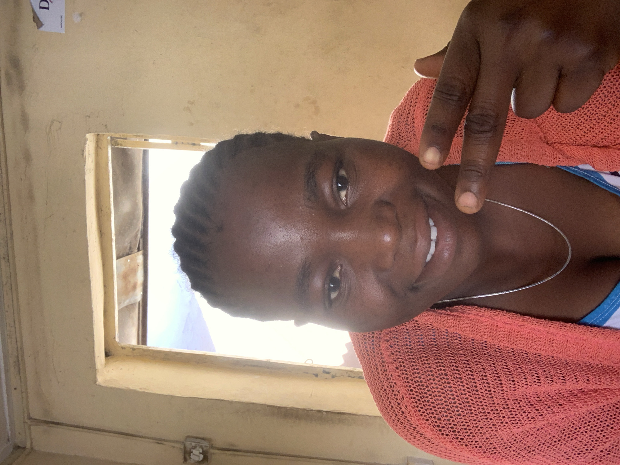 A portrait of a young woman with dark skin and braided hair. She is smiling and making a "peace" sign with her right hand. She is wearing a silver necklace and a coral-colored sweater.
The background is a yellowish wall with a window. The window is old, with peeling paint, and the view is of the outside.
The image was taken indoors, likely in a residential or office space in Jos, Nigeria.