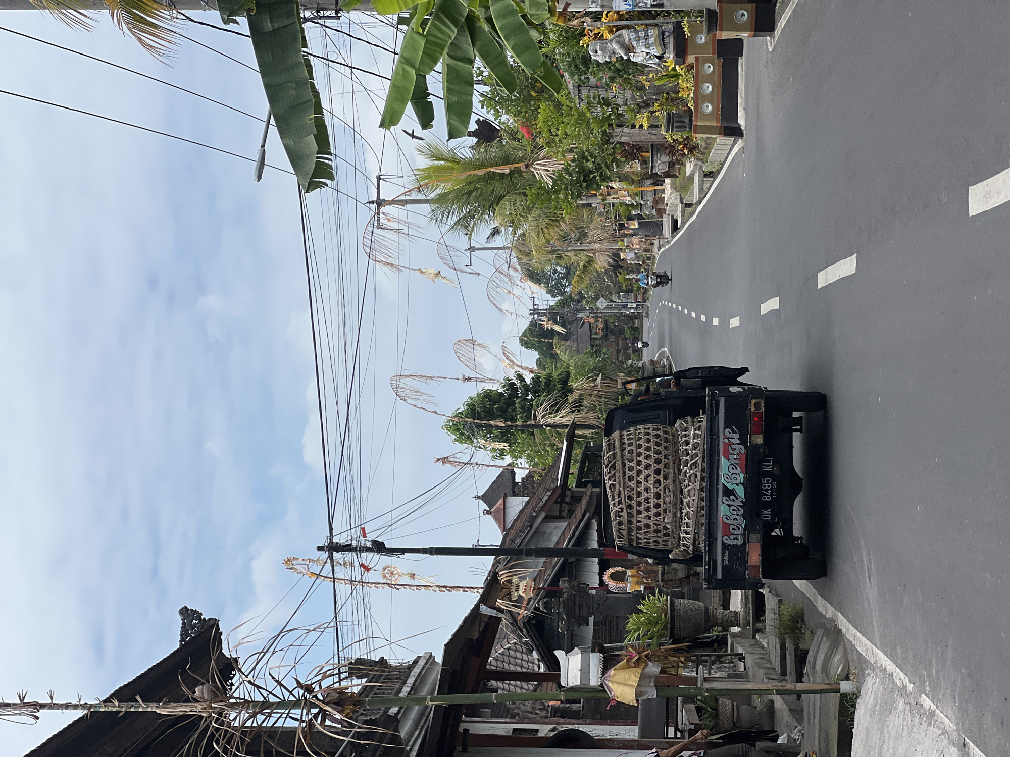 A black pickup truck is parked on the right side of a paved road in Sukawati, Indonesia. The truck bed is filled with several woven baskets. The license plate reads "DK 8485 KL". The words "bebek bengil" are written in blue cursive on the tailgate. The truck is facing away from the viewer. The road is lined with buildings and vegetation, and overhead power lines are visible. Two people are visible near the truck, one standing and one partially obscured by the truck. In the distance, a motorcycle is being ridden by a person wearing a blue shirt. The sky is cloudy.